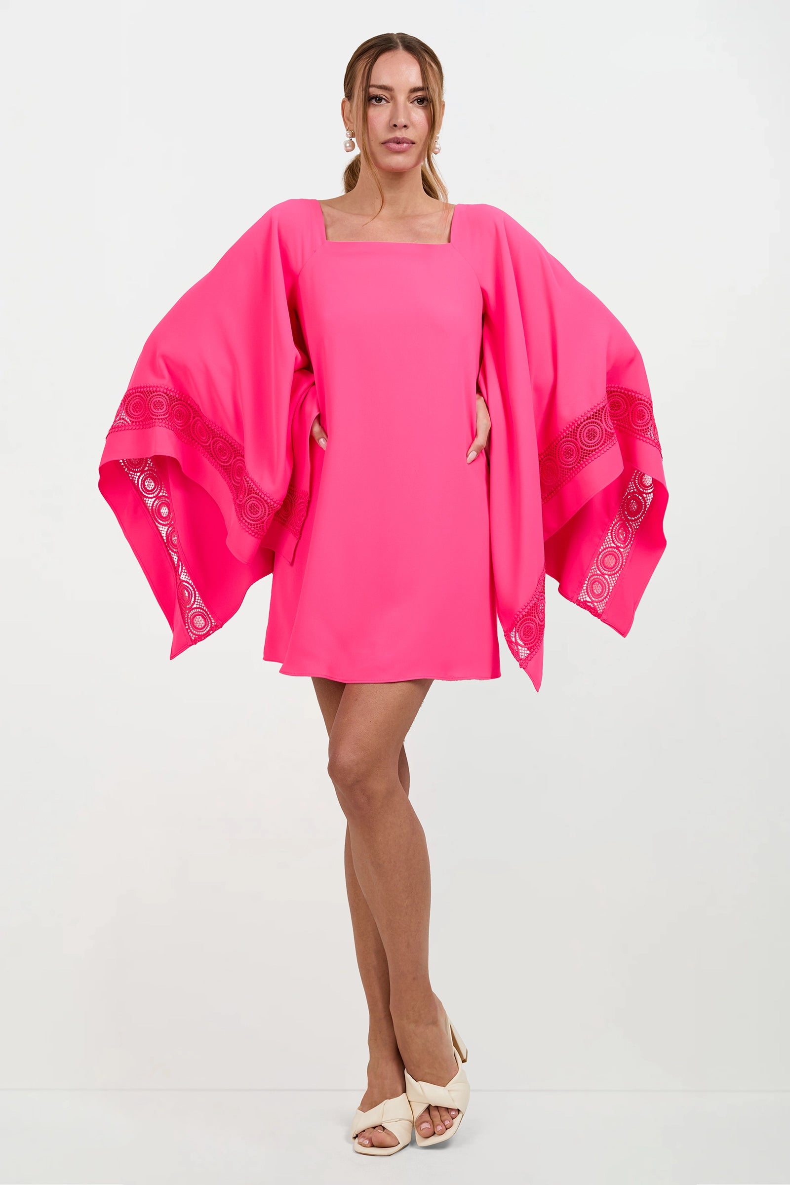 A woman poses against a plain background wearing the Aqualane Side Flounce Dress, a bright pink mini dress with wide, flowing sleeves and lace-like trim, paired with white open-toe heels and one leg crossed over the other.