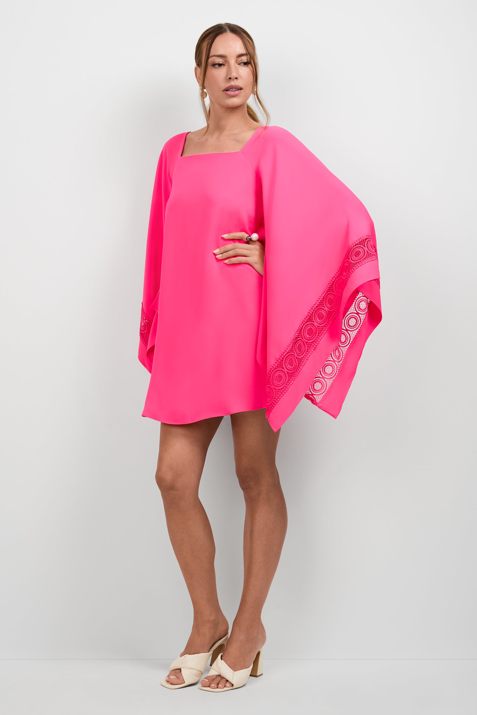 A woman stands against a plain background wearing the Aqualane Side Flounce Dress, a bright pink, loose-fitting style with wide flowing sleeves and decorative trim, paired with cream-colored heeled sandals and her hair tied back.