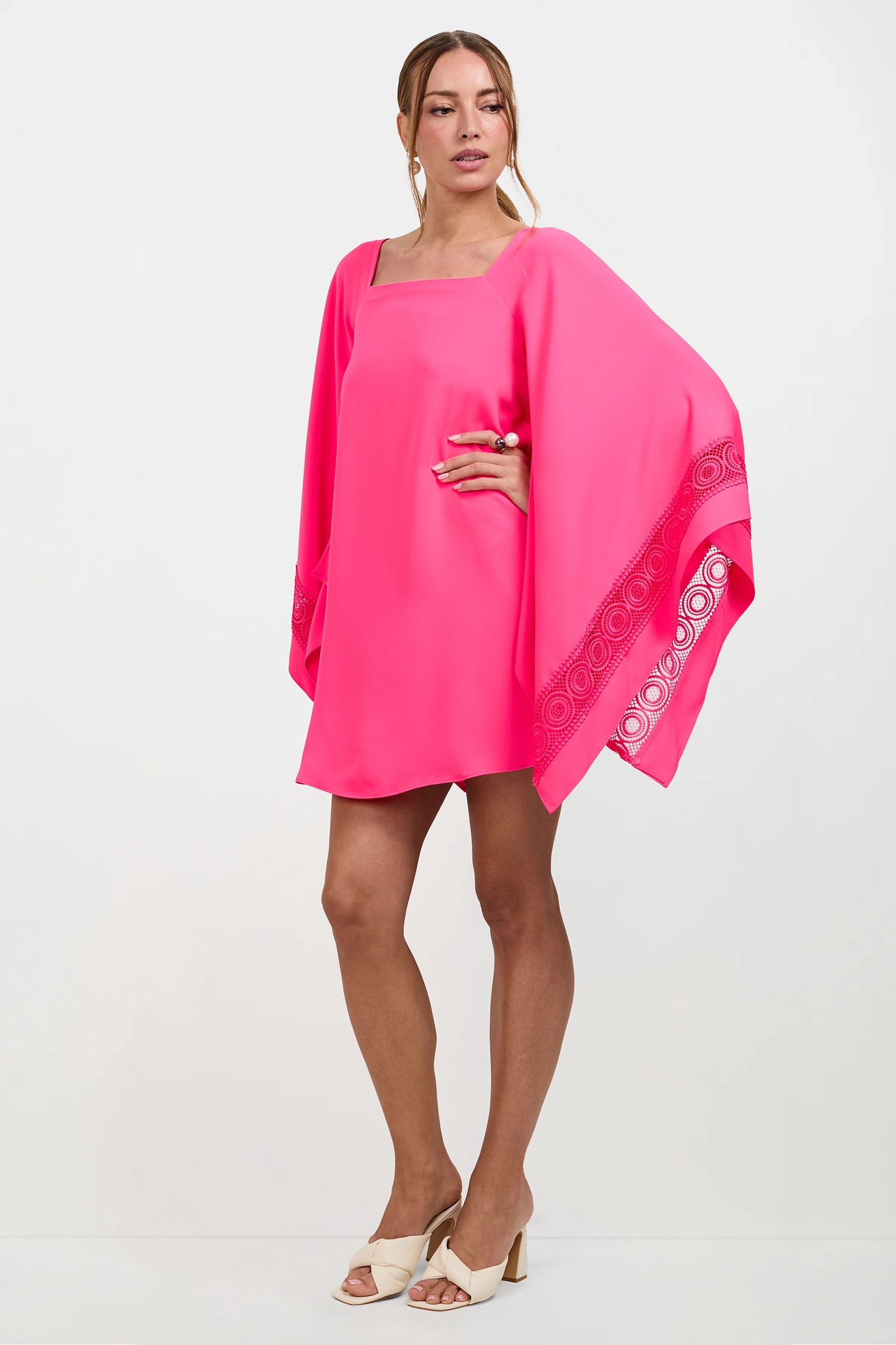 A woman poses against a white background in the Aqualane Side Flounce Dress in Magenta Milano with wide, patterned sleeves, one hand on her hip, and open-toed cream heels.