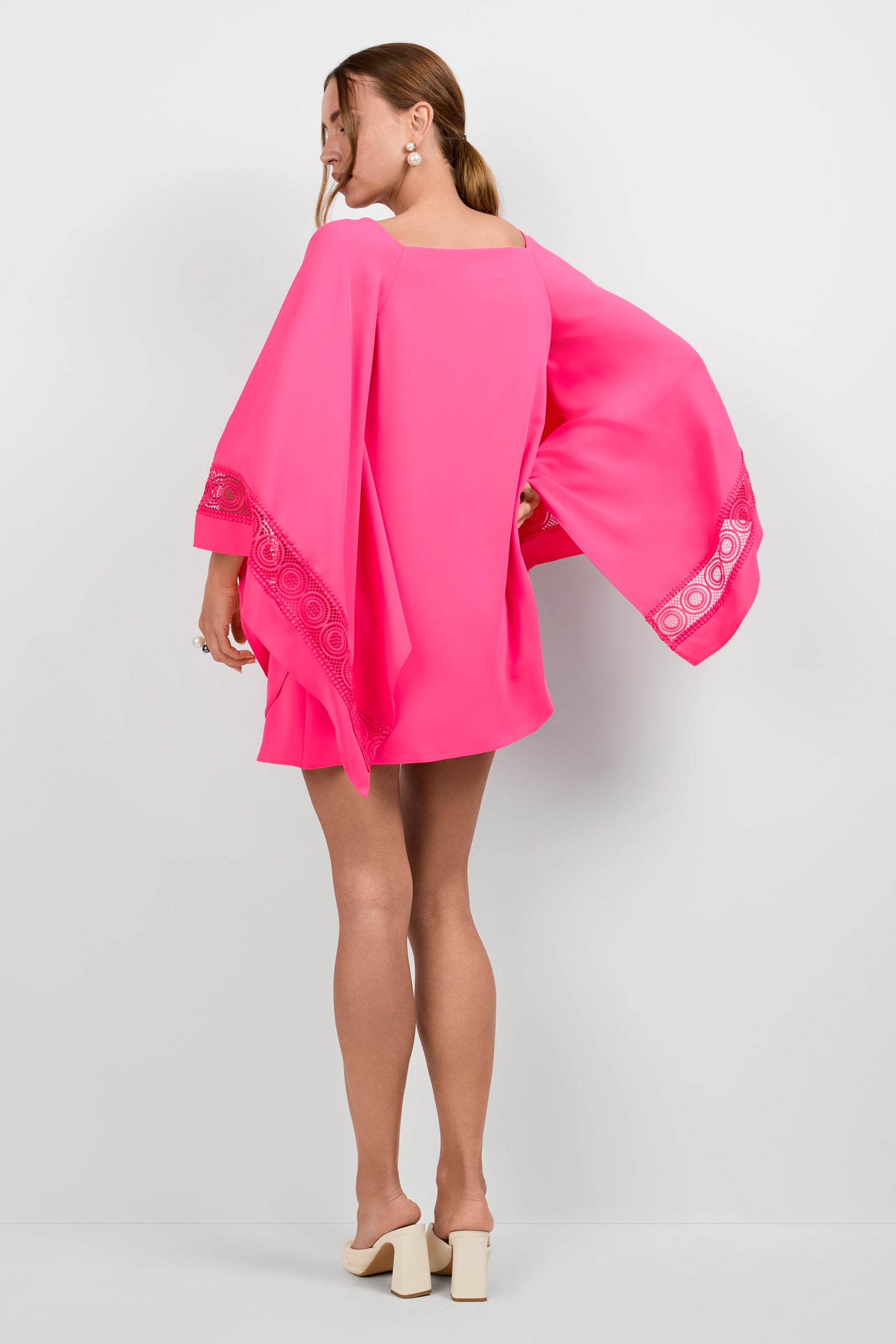 A woman, hair tied back, models the Aqualane Side Flounce Dress—a bright pink mini dress with wide, flowing sleeves and decorative trim—paired with beige block-heeled shoes against a plain white background.
