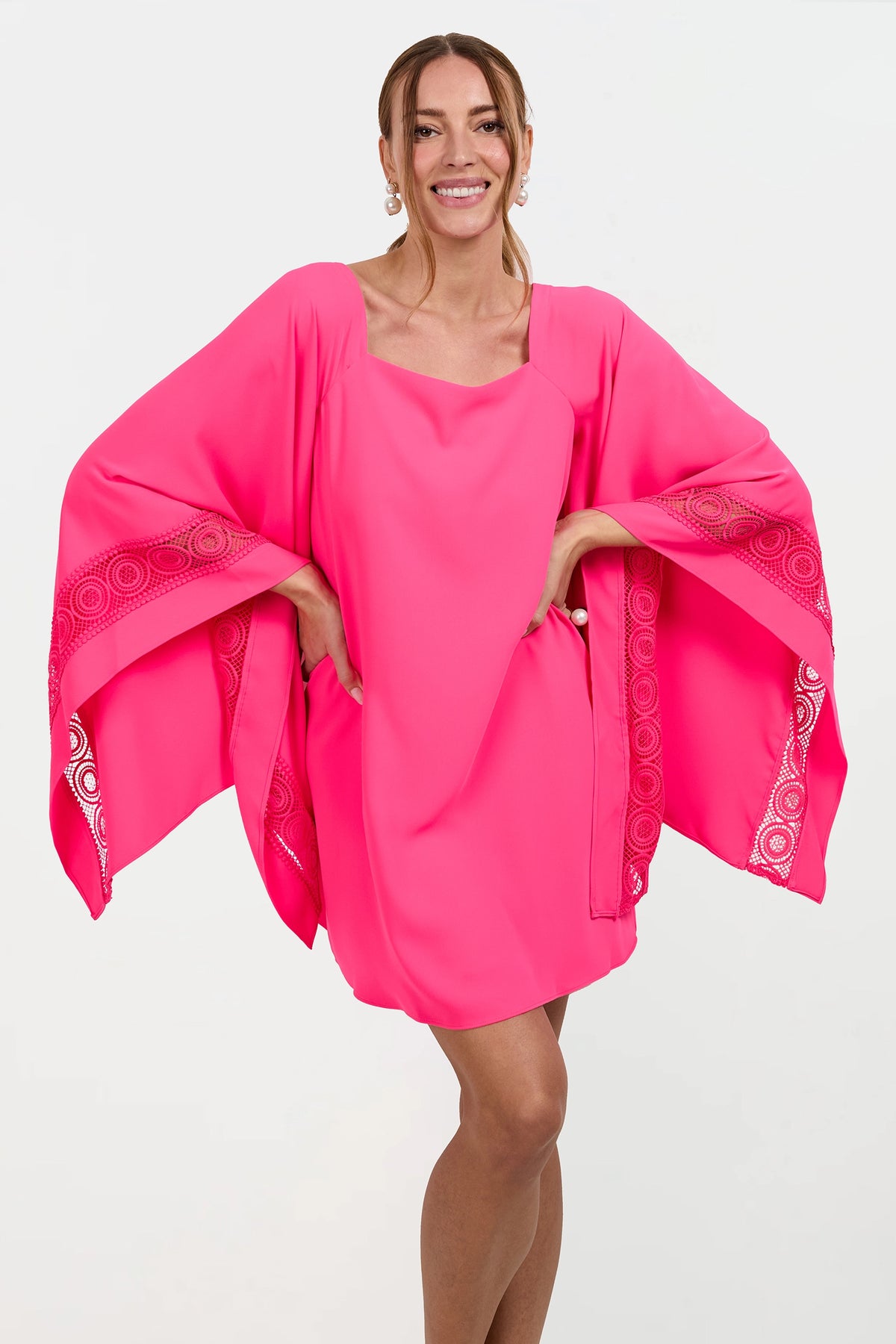 A woman smiles in the Aqualane Side Flounce Dress, a bright pink outfit with wide, flowing sleeves adorned with circular lace patterns. She stands confidently with her hands on her hips against a plain white background.