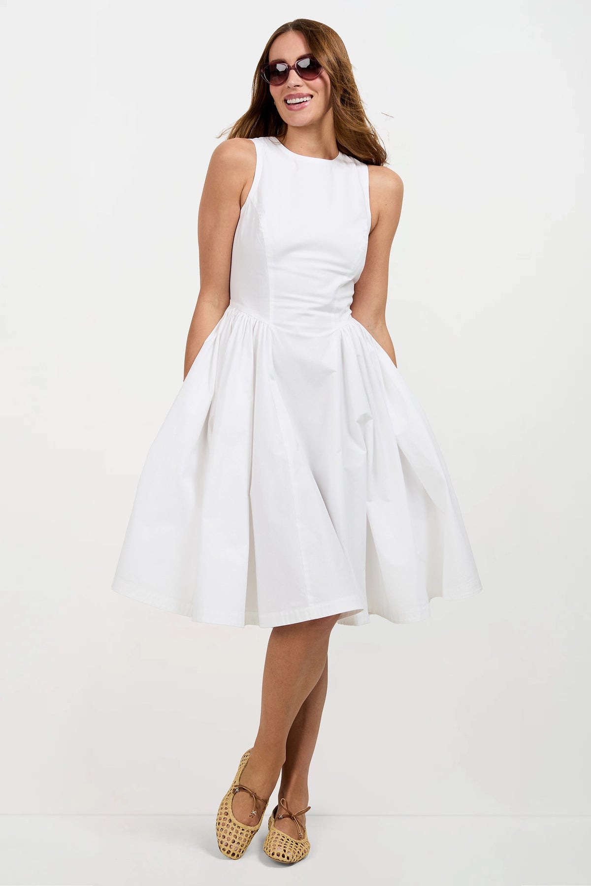 A woman wearing the Lombardy Midi Dress, a sleeveless white knee-length dress, along with sunglasses and tan woven shoes, stands smiling with her arms relaxed at her sides against a plain white background.