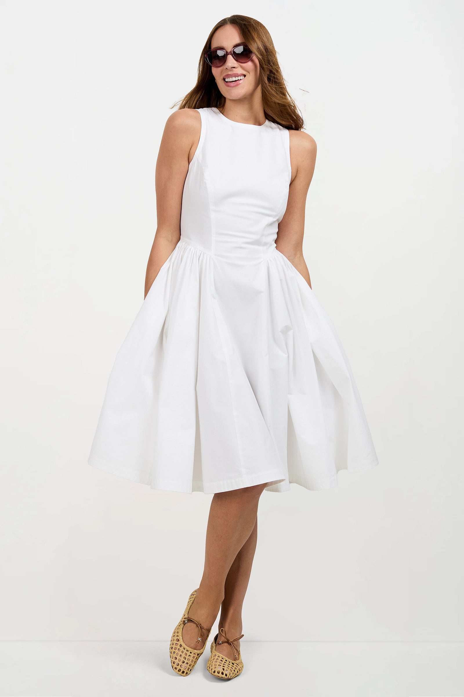 A woman wearing the Lombardy Midi Dress, a sleeveless white knee-length dress, along with sunglasses and tan woven shoes, stands smiling with her arms relaxed at her sides against a plain white background.