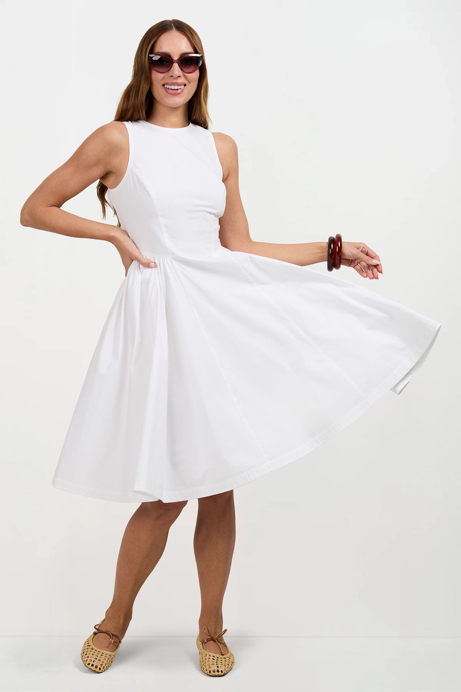 A woman poses playfully against a plain white background, wearing the Lombardy Midi Dress, sunglasses, tan woven shoes, and red bangles while holding the skirt of her dress to the side.