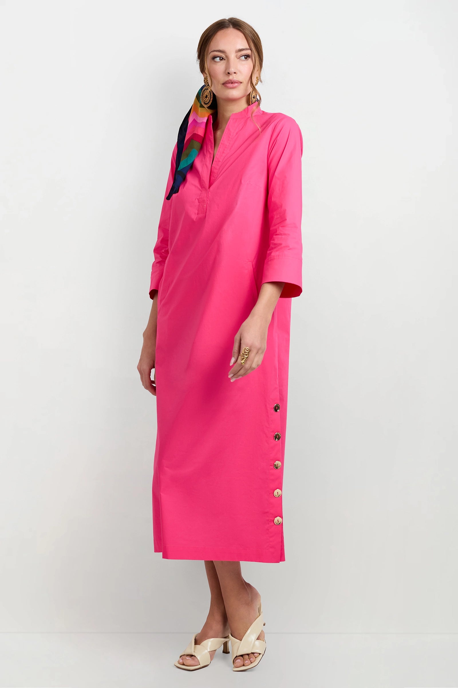 She wears the Capella Mandarin Collar Maxi Dress in bright pink with beige heeled sandals, gold earrings and rings, and a colorful scarf tied in her hair.