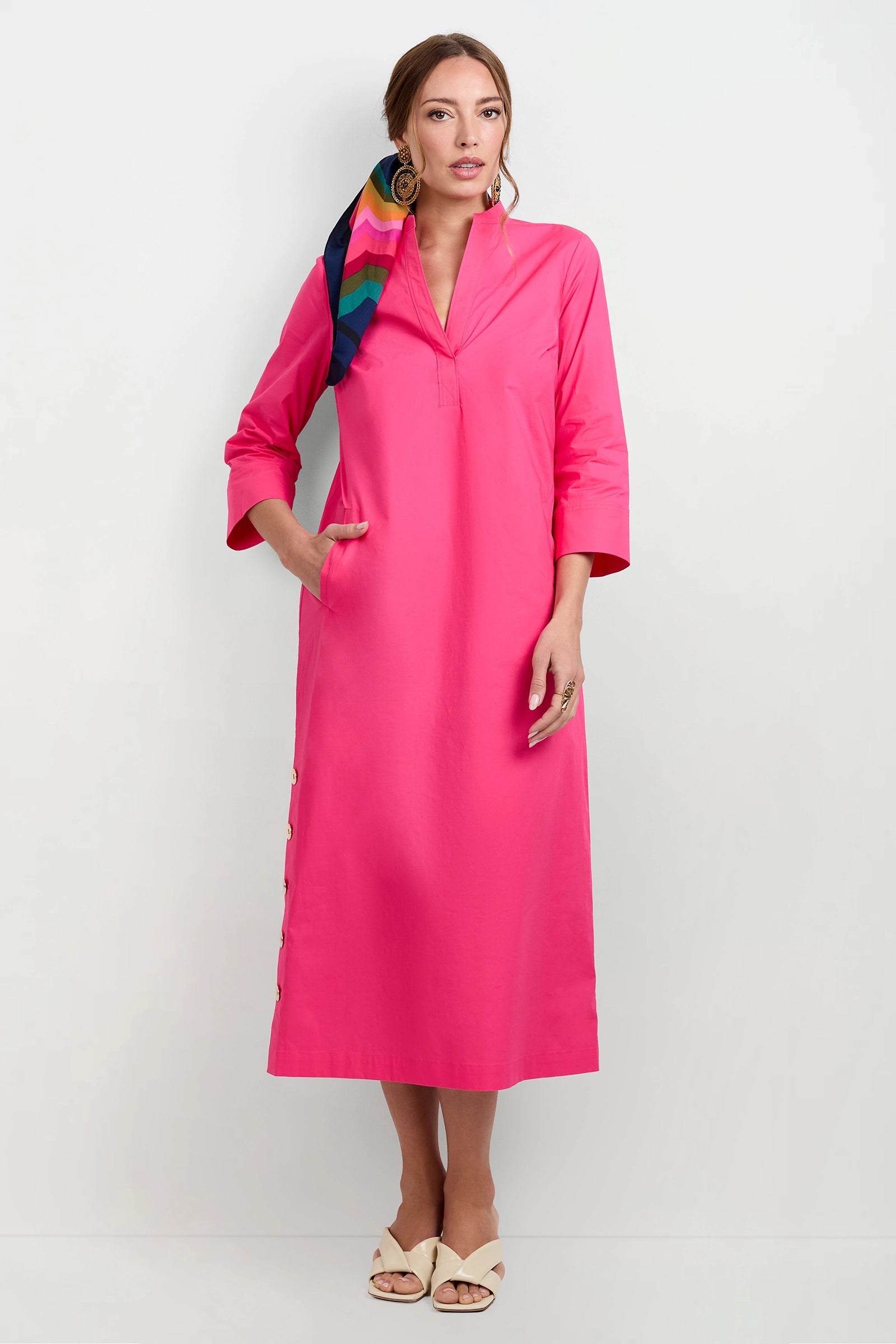 A woman stands against a plain white background wearing the Capella Mandarin Collar Maxi Dress in Magenta Milano, beige open-toe shoes, gold hoop earrings, and a multicolored scarf in her hair.