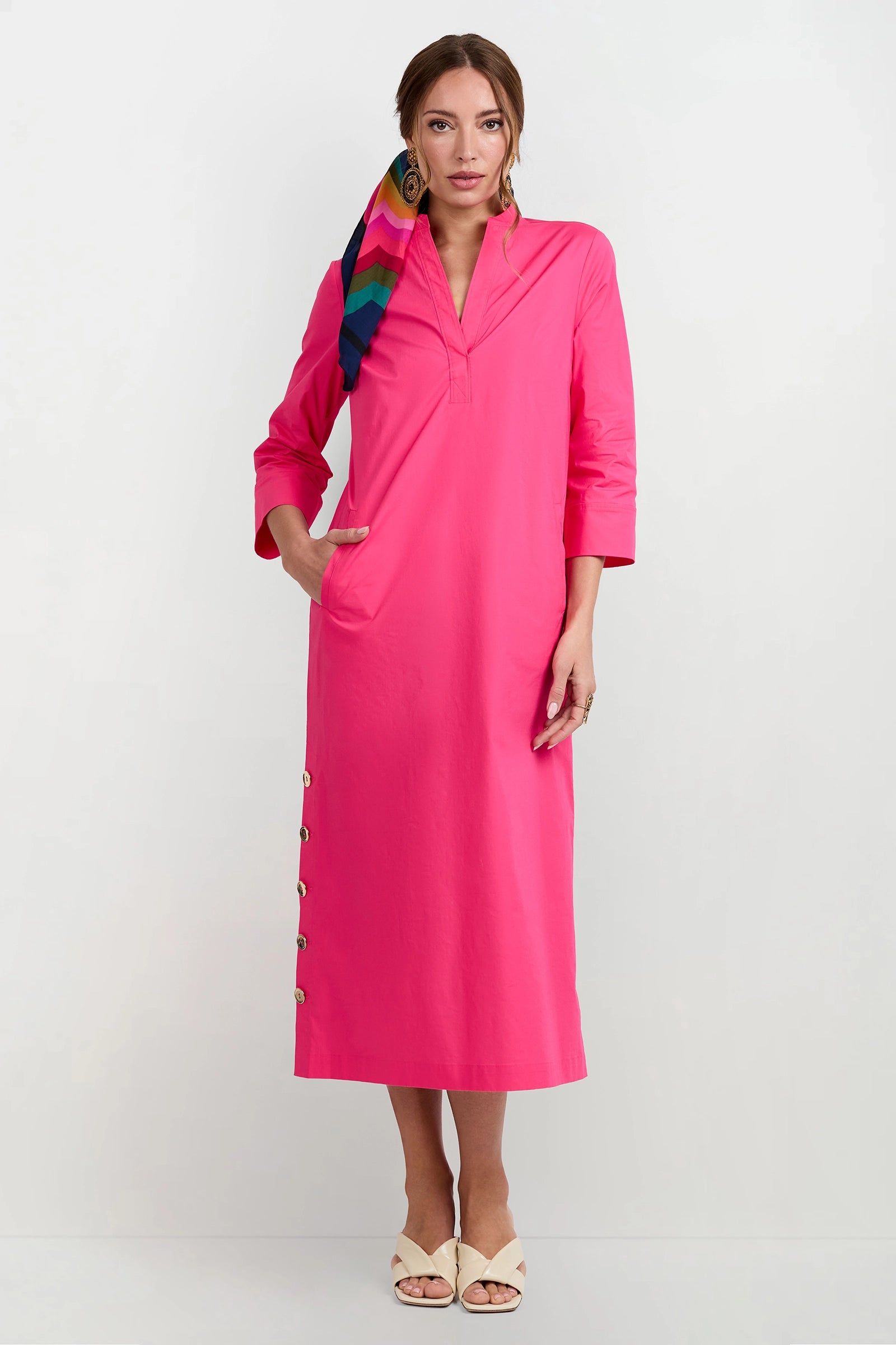 A woman poses against a white background in the Capella Mandarin Collar Maxi Dress, a bright pink, long-sleeve piece with side hem buttons. She wears beige sandals, large earrings, and a colorful striped headscarf in her hair.