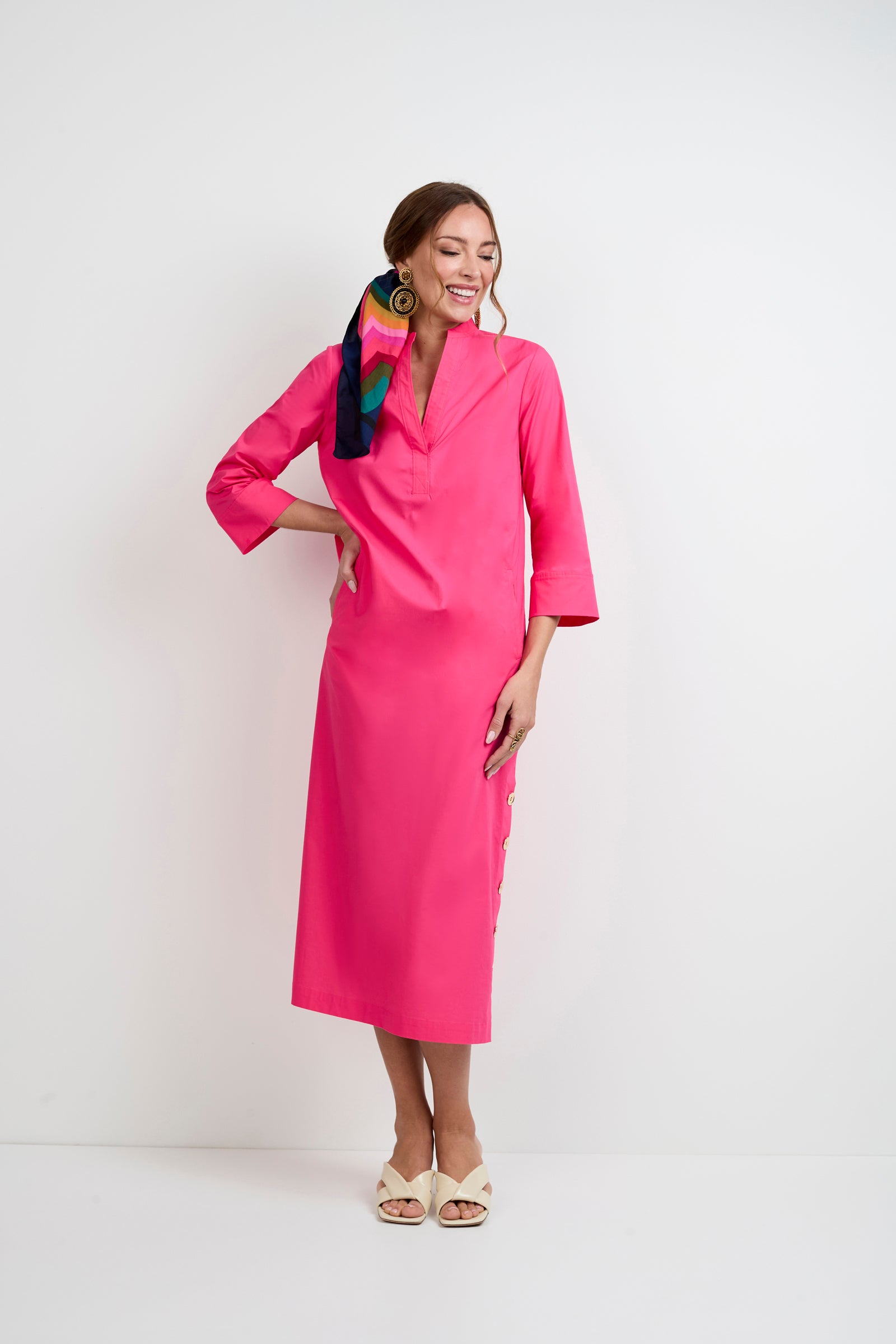 A woman stands against a white background wearing the Capella Mandarin Collar Maxi Dress. She has a colorful scarf in her hair, large earrings, and white open-toe shoes, smiling as she looks to the side.