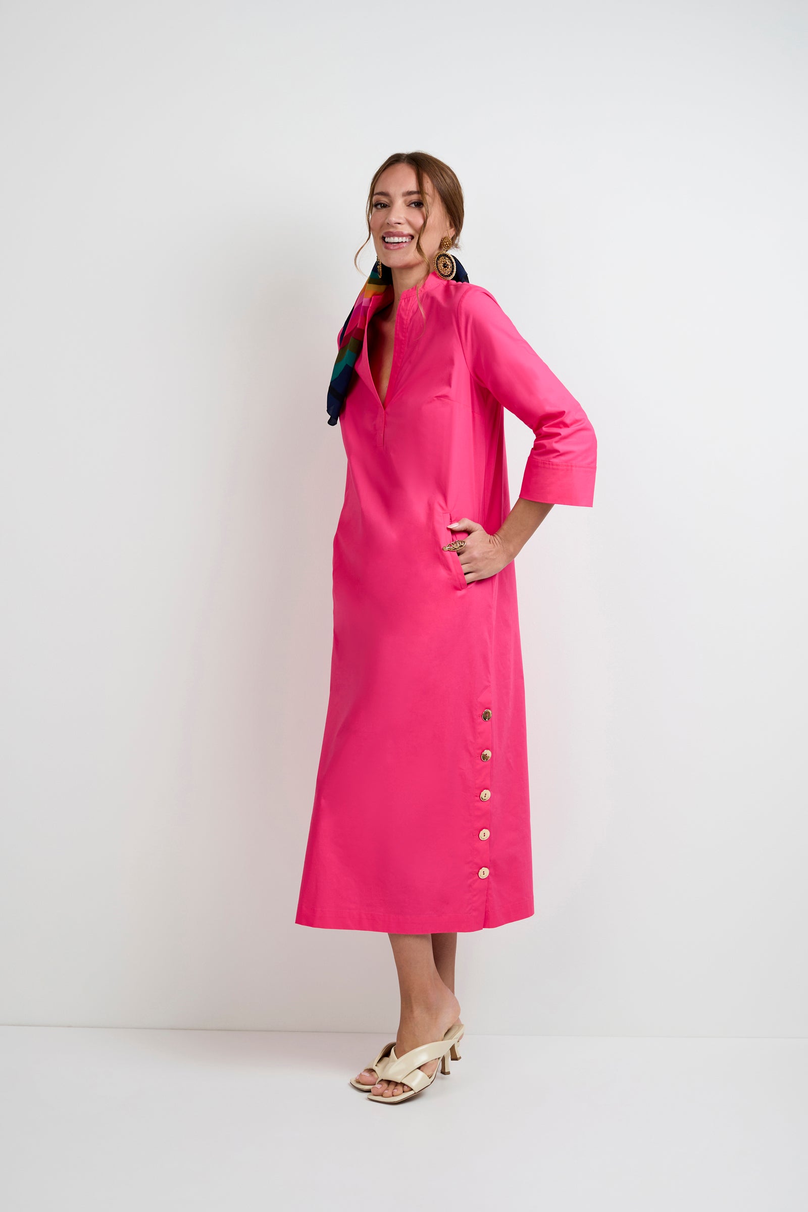 A woman poses against a white background in the Capella Mandarin Collar Maxi Dress, featuring a bright pink hue, three-quarter sleeves, side buttons, and cream heeled sandals. She completes the look with bold earrings and a colorful hair scarf.