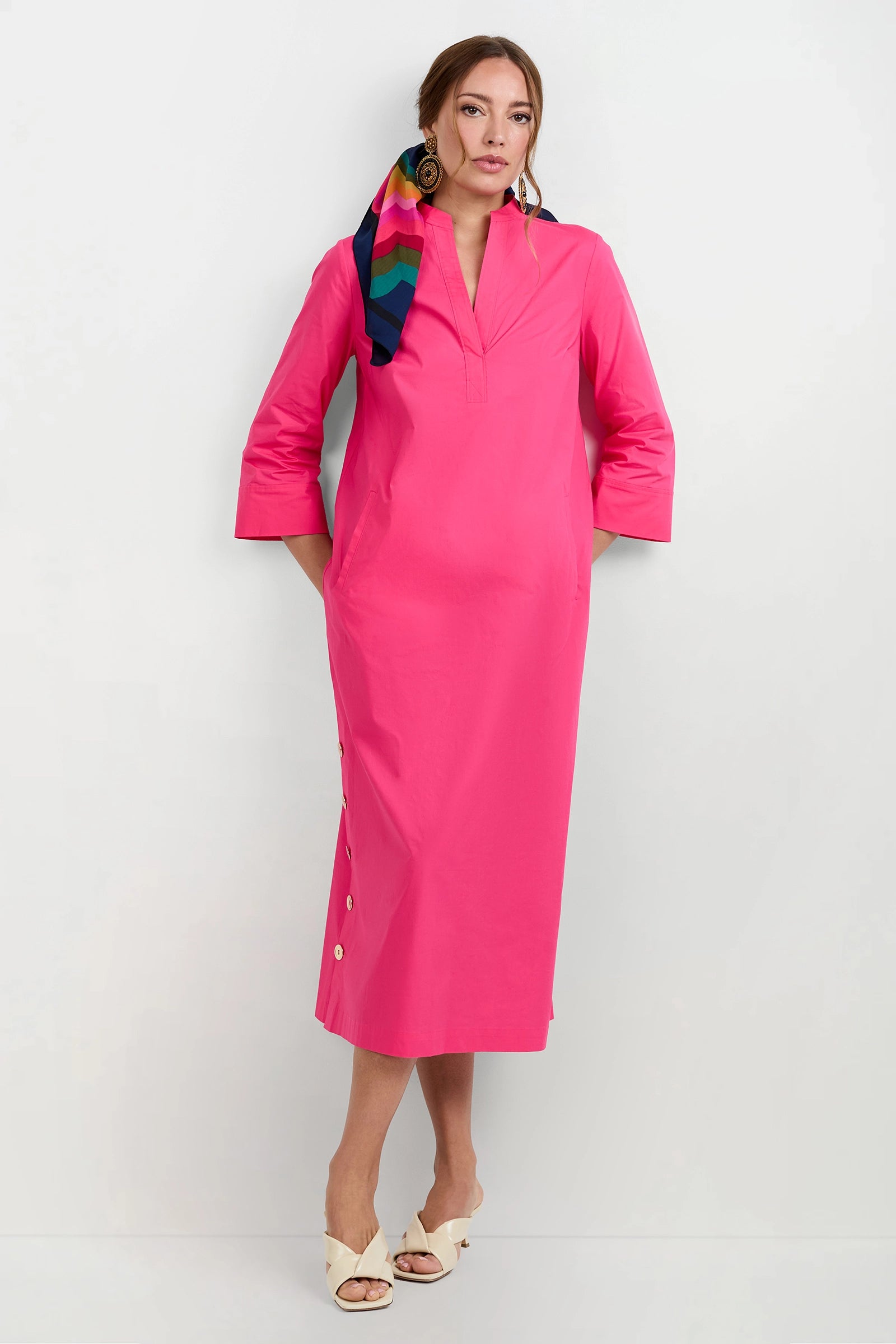 A woman wears the Capella Mandarin Collar Maxi Dress—bright pink, long-sleeved, midi length with side buttons—styled with a colorful hair scarf, large earrings, and beige heeled sandals against a plain white background.