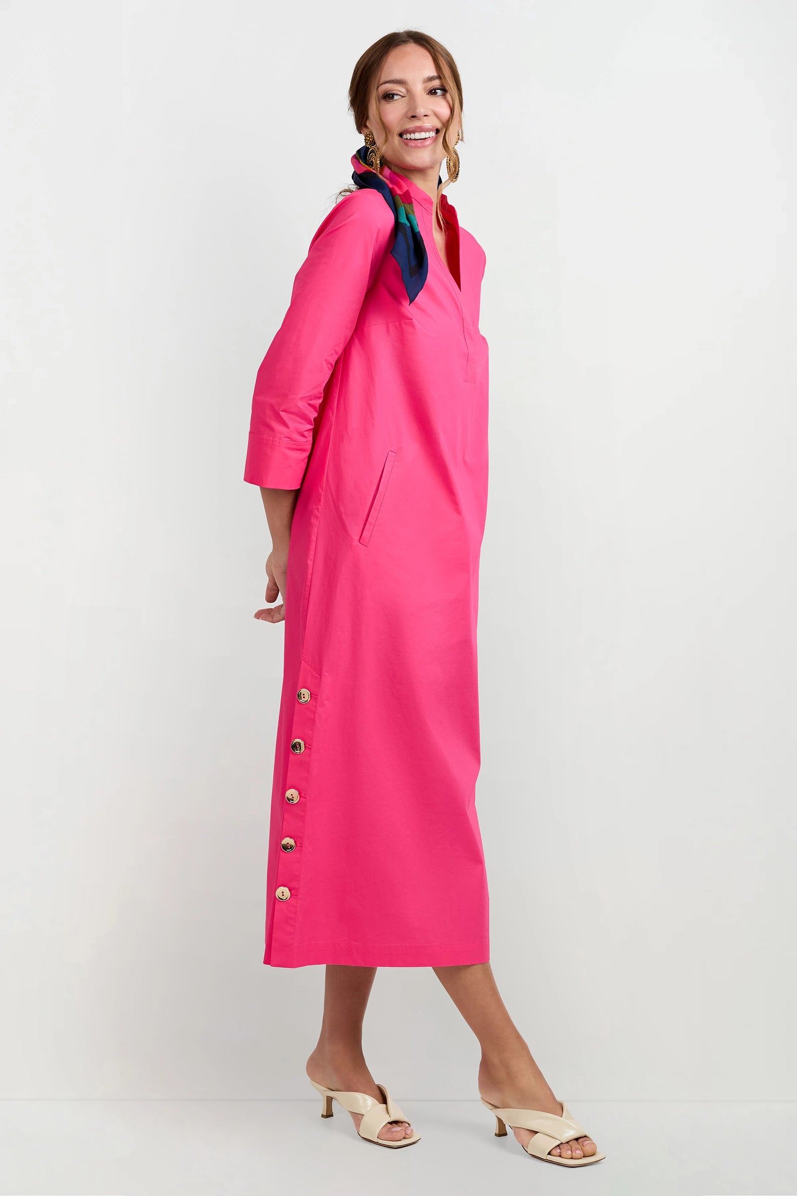 A smiling woman models the Capella Mandarin Collar Maxi Dress, a bright pink, long-sleeve maxi with side button details. She pairs it with cream heeled sandals and a navy-pink scarf, posing against a plain white background.