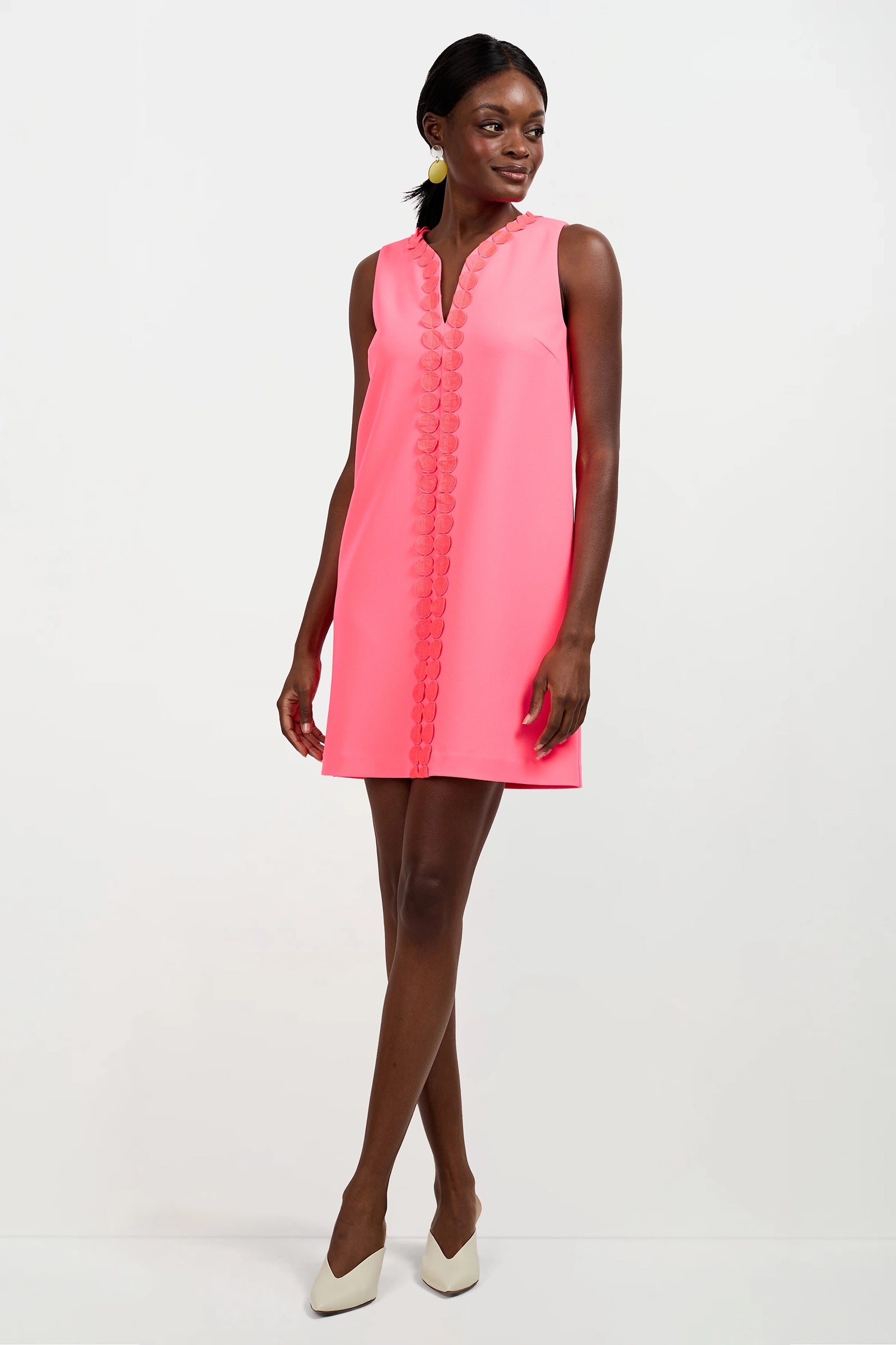 A woman stands against a plain white background wearing the Pavia Mini Dress—sleeveless, bright pink with scalloped front trim—paired with white heels.