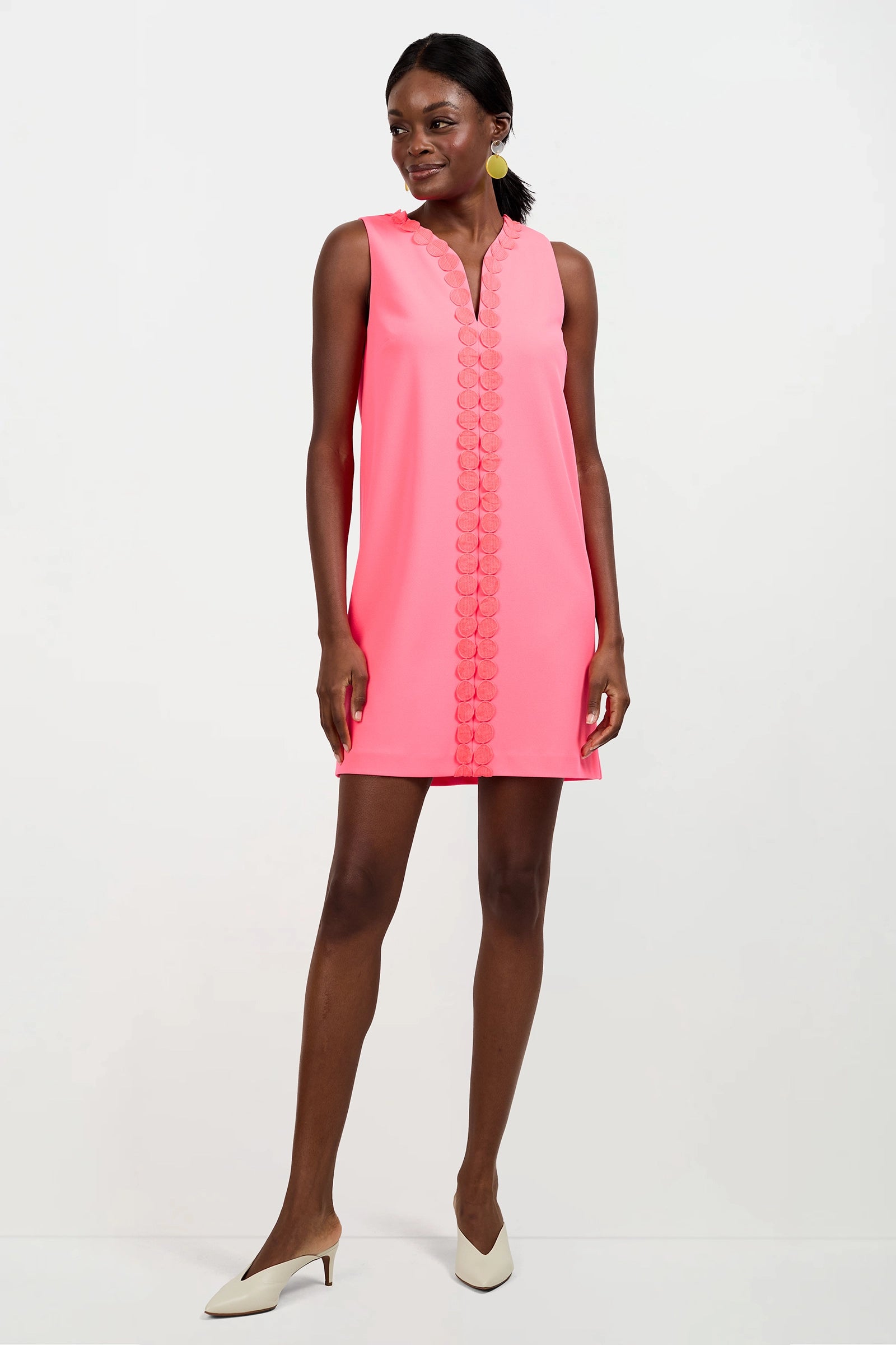 A woman in the Pavia Mini Dress—a sleeveless, bright pink shift with scalloped center trim—stands against a plain white background wearing white heels.