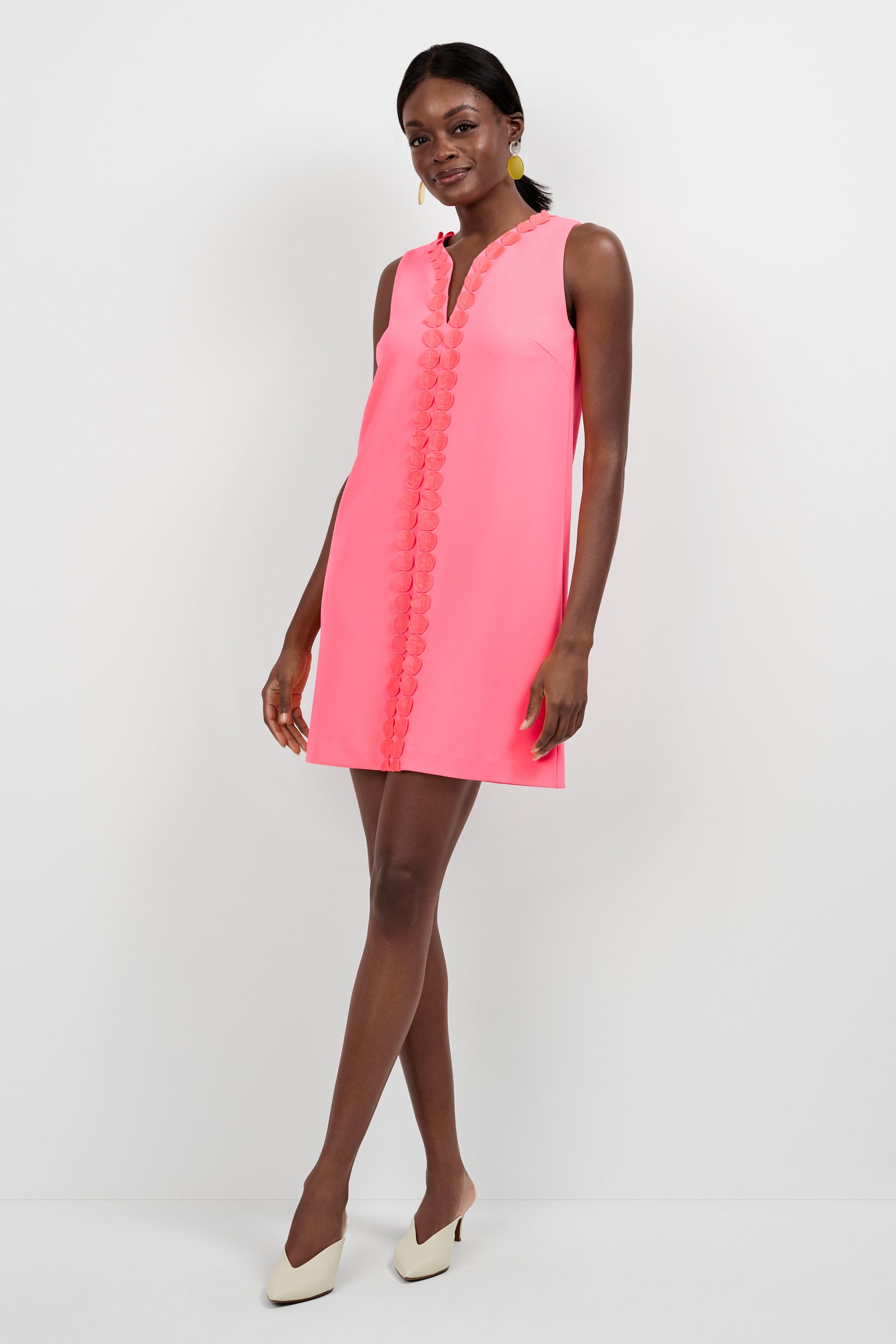 A woman stands against a white background, wearing the Pavia Mini Dress in Corallo with textured center trim and off-white heels. She smiles softly, one hand slightly lifted.