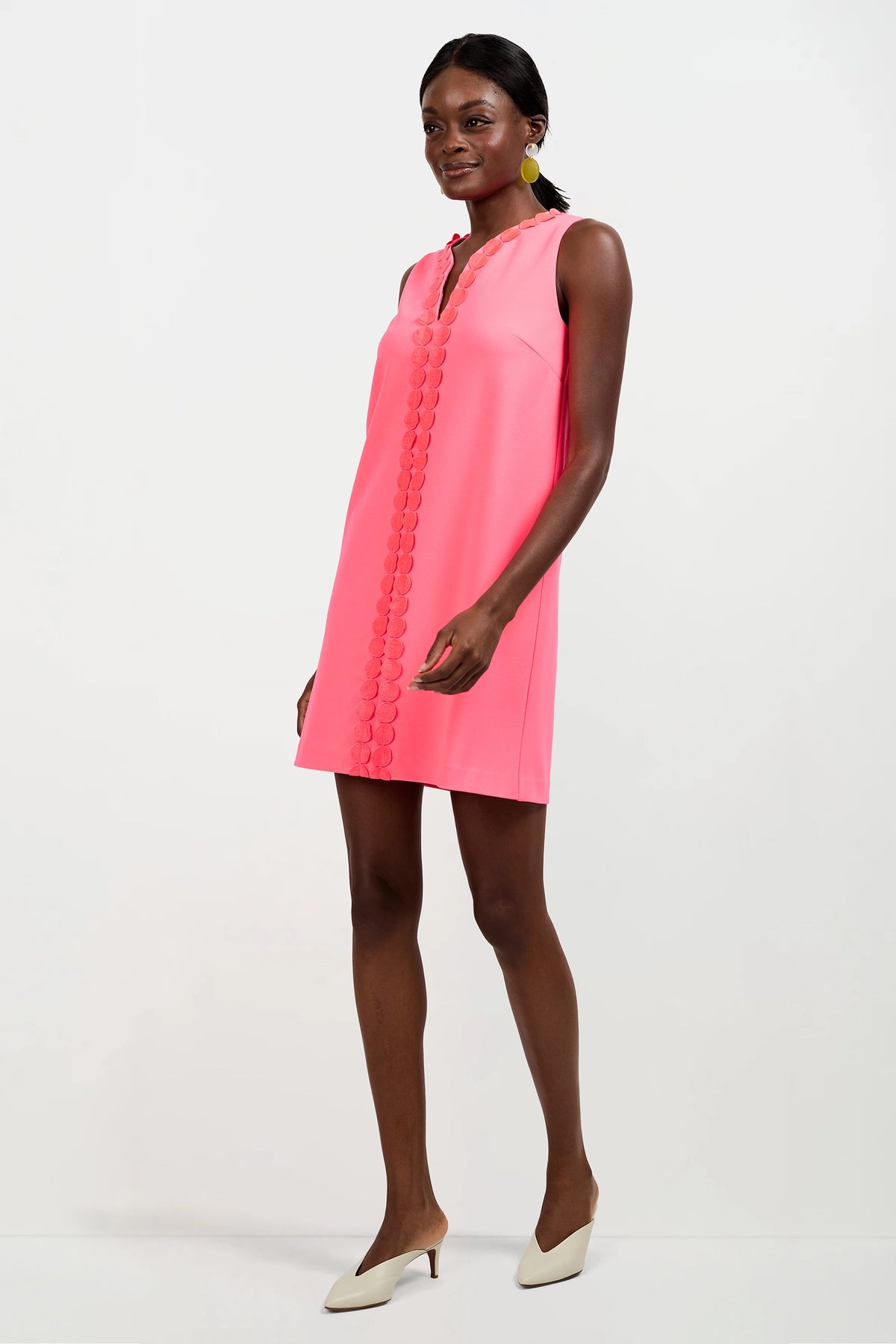 A woman wears the Pavia Mini Dress, a sleeveless, knee-length pink dress with scalloped front trim. She pairs it with beige heels and yellow statement earrings, standing against a plain white background.
