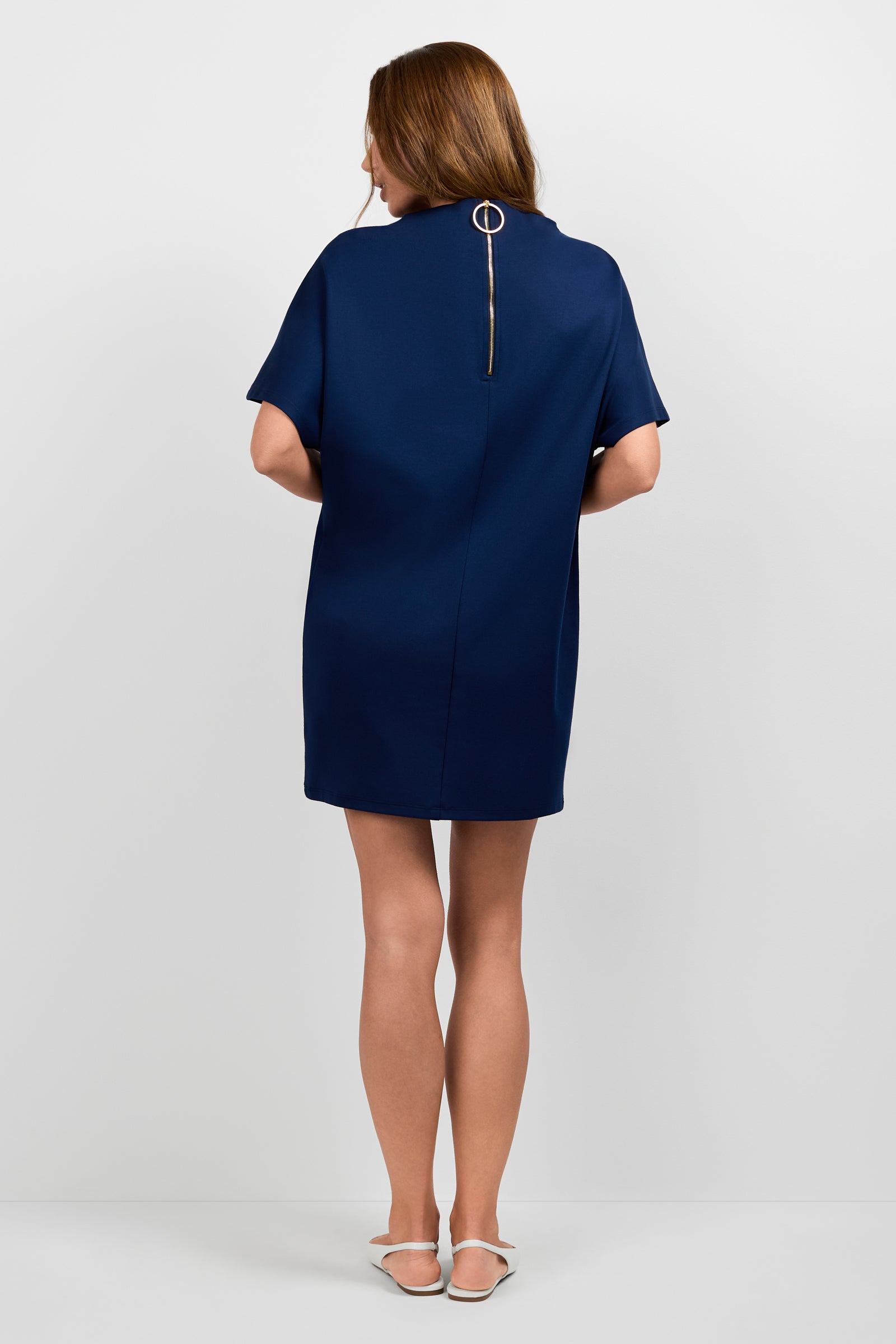 A woman with light brown hair faces away, wearing the Storica Mini Dress in dark blue with short sleeves and a back zipper with a ring pull. She pairs it with white flats and stands against a plain background.