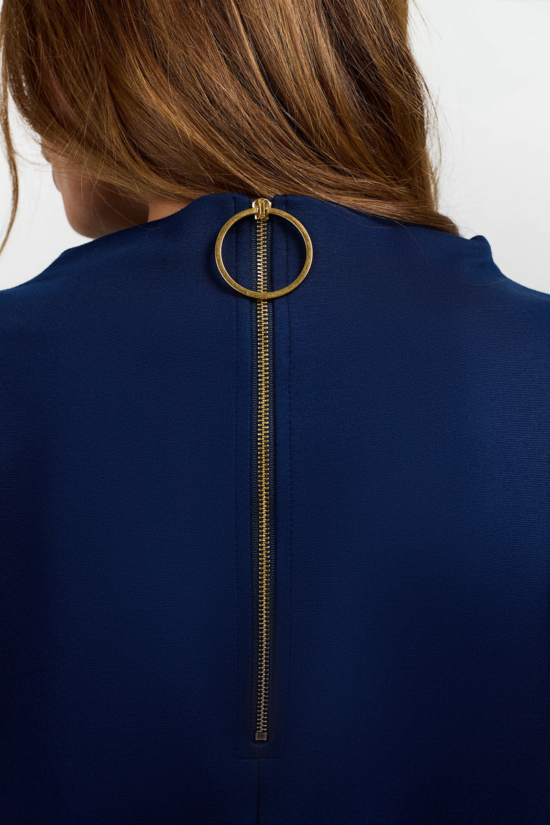 A woman with brown hair is shown from behind wearing the Storica Mini Dress in dark blue, featuring a visible gold zipper with a large circular pull tab at the back of the neck.