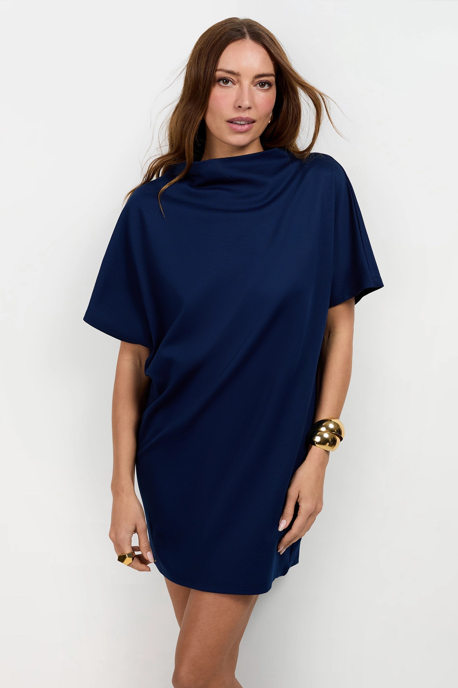 A woman with long brown hair wears the Storica Mini Dress, a loose-fitting, short-sleeved navy blue dress with a high neckline. She accessorizes with chunky gold bracelets and a gold ring, posing against a plain light background.