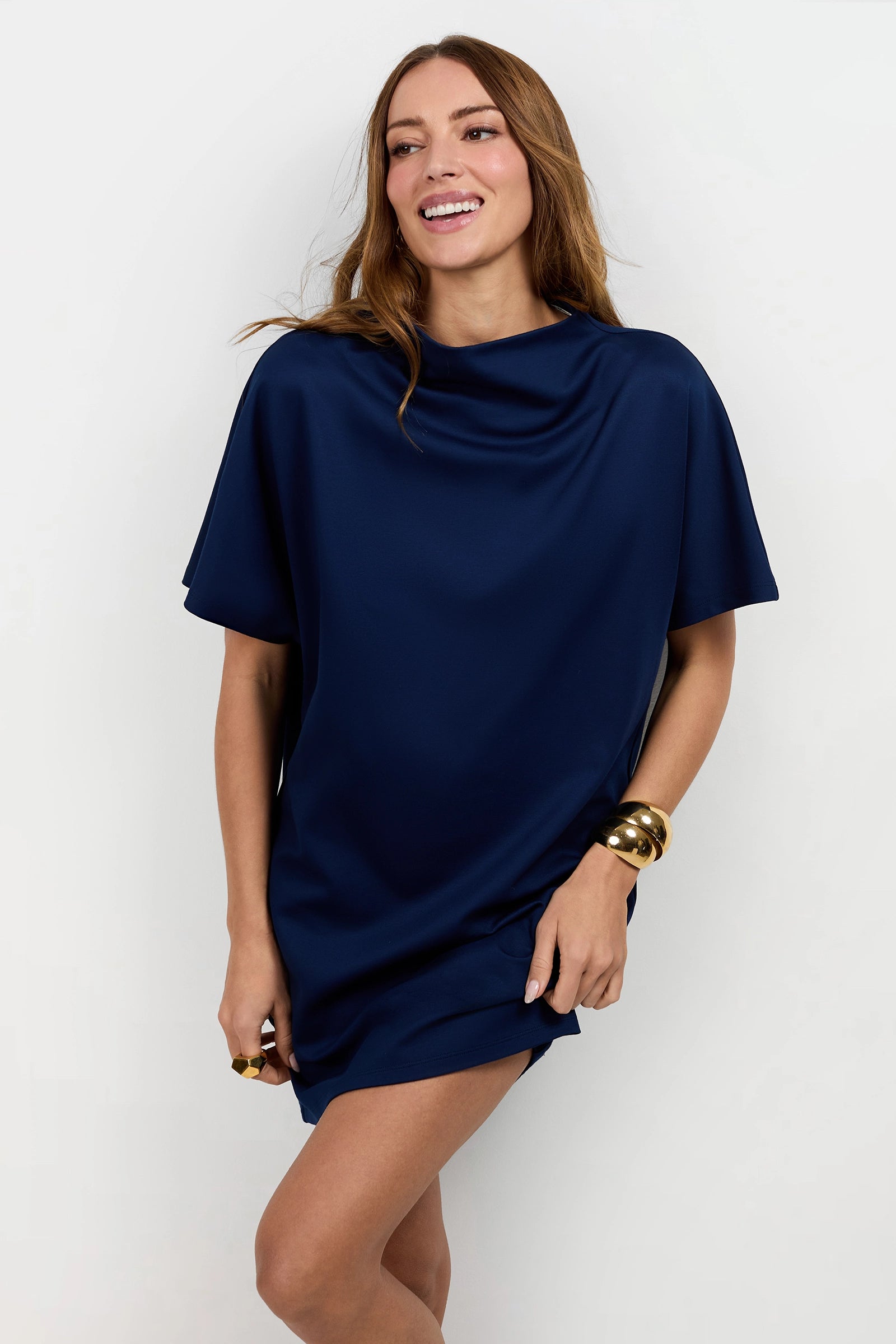 A woman with long brown hair smiles in the Storica Mini Dress—a loose-fitting, short navy blue dress with short sleeves—styled with large gold bangles, standing against a plain white background.
