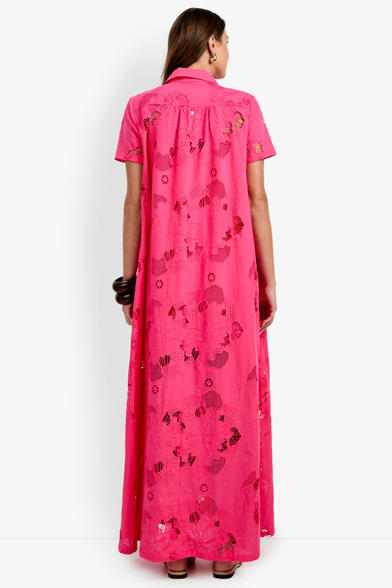 A woman stands facing away, wearing the Kon Tiki Maxi Dress—a bright pink, floor-length dress with short sleeves and floral cut-out patterns throughout the fabric.