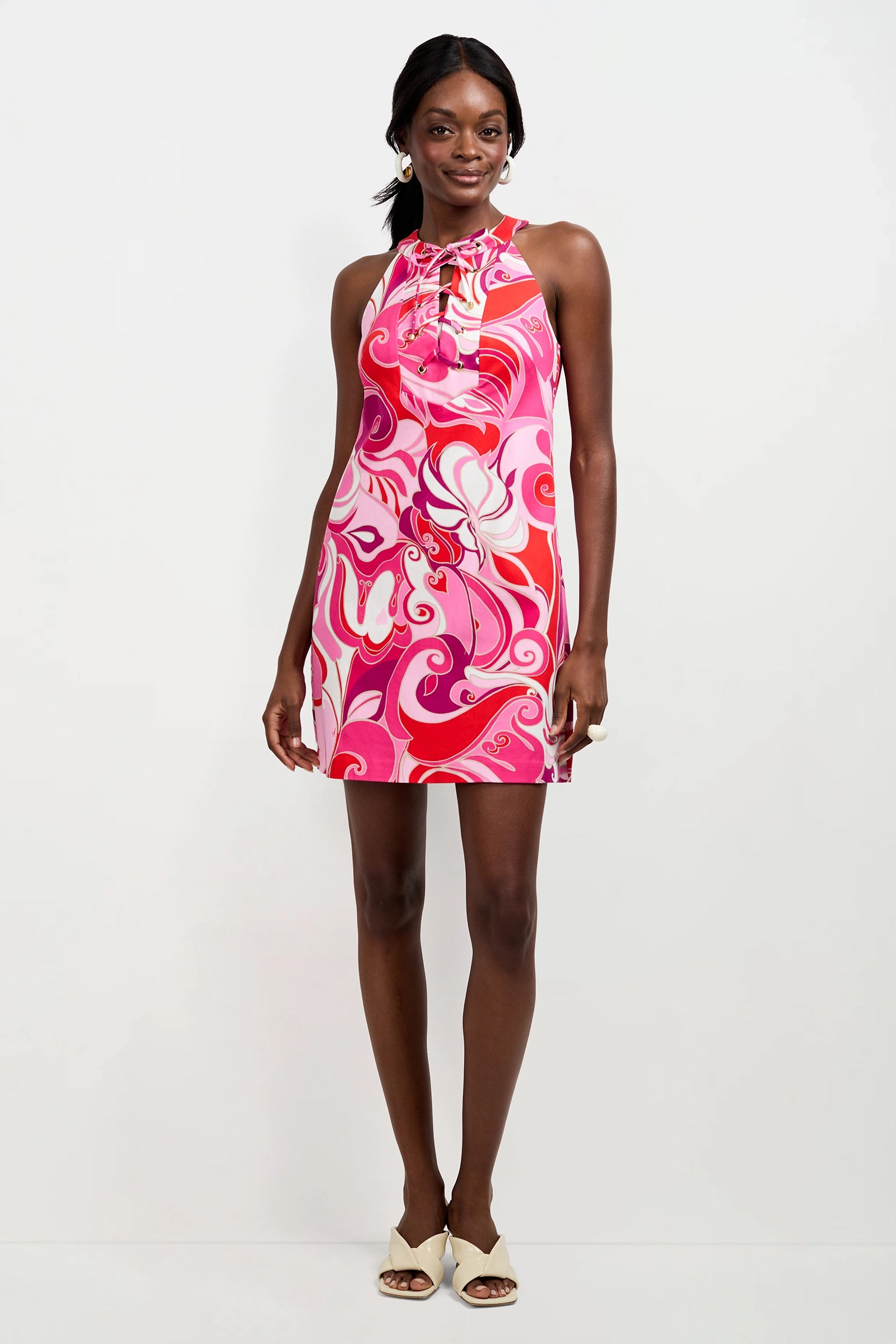 A woman poses against a white background wearing the Cappuccia Lace-Up Mini Dress in Darsena, featuring a sleeveless halter neck and bold swirl pattern, paired with cream open-toe heels.