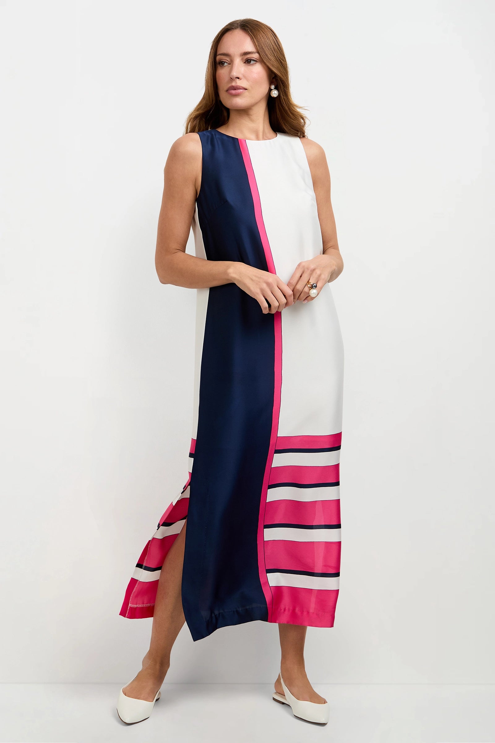 A woman wearing the Moderna Linee Midi Dress—sleeveless, ankle-length, with a navy vertical panel, white side, bold pink stripes at the hem—paired with white flats, stands against a plain white background.