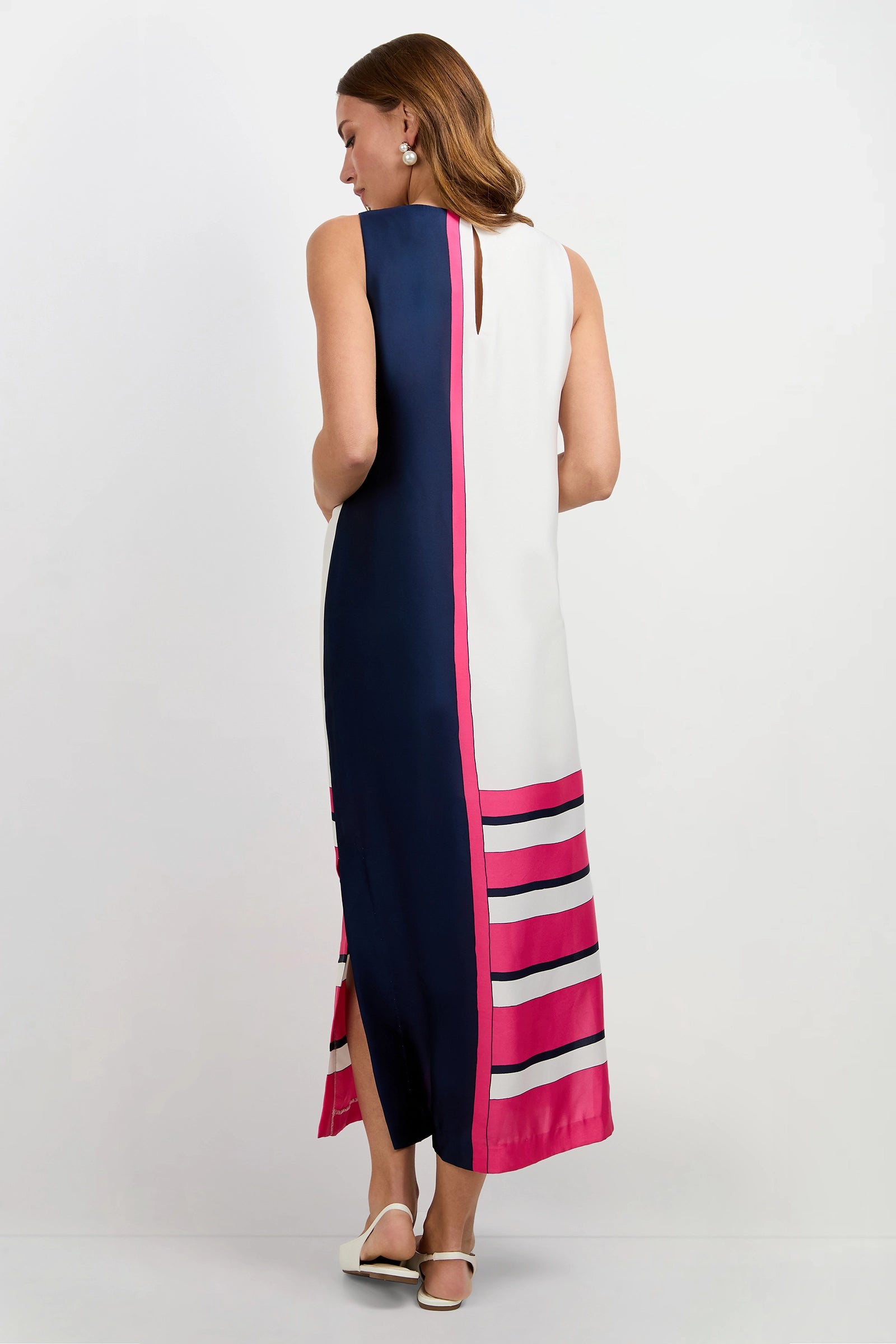 A woman with shoulder-length brown hair, in white sandals, faces away wearing the Moderna Linee Midi Dress—a sleeveless, ankle-length style featuring a navy and white vertical split and pink-magenta horizontal stripes near the hem.