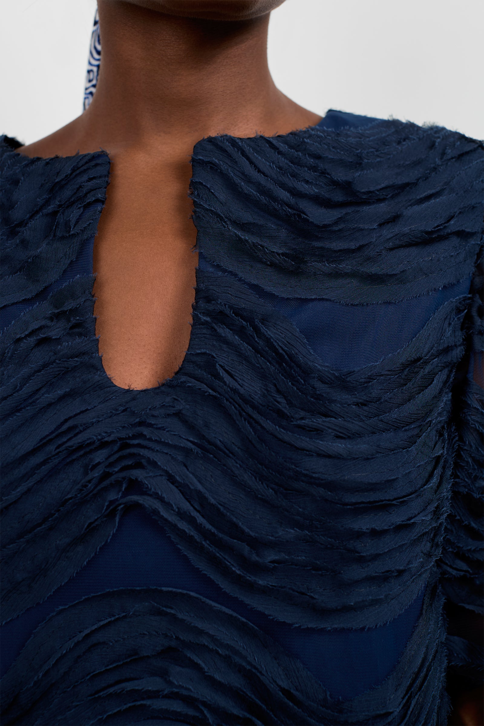 A close-up of a person wearing the Torchino Organza Mini Dress, featuring textured navy blue fabric with layered, wavy patterns and a small V-shaped neckline. The background is plain and light-colored.