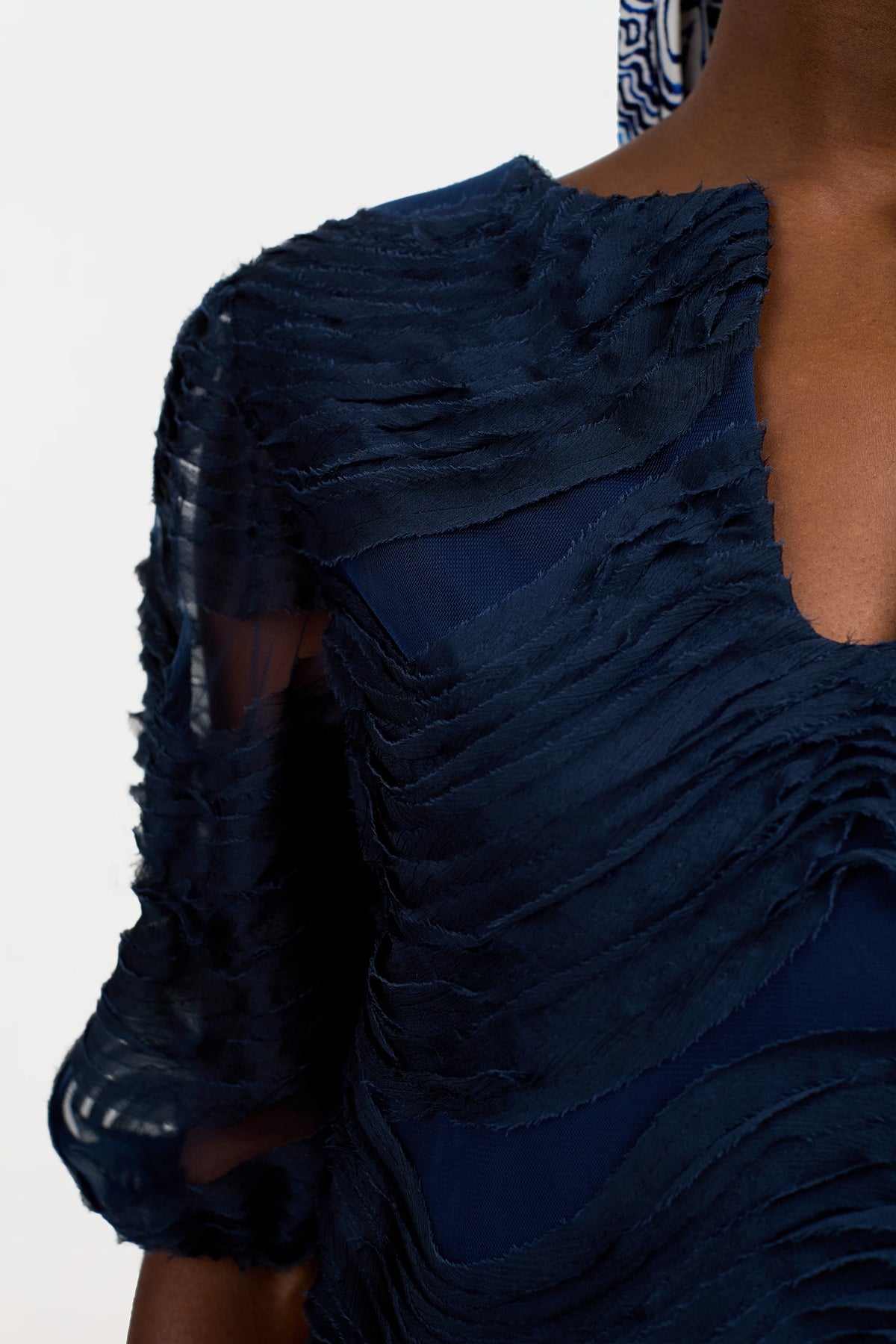 Close-up of a person wearing the Torchino Organza Mini Dress in navy blue, featuring layered sheer fabric, a deep V-neckline, horizontal ruffles, and semi-transparent sleeves. Only part of the upper body is visible.