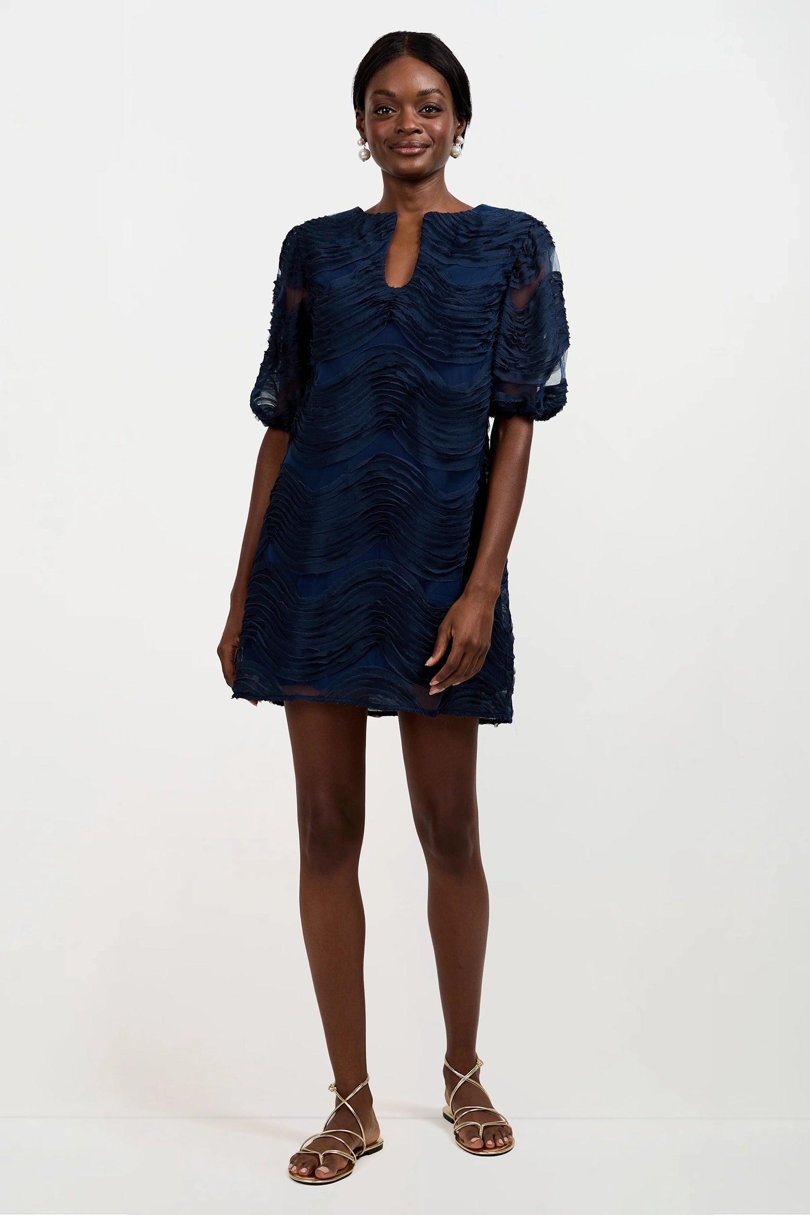 A woman stands against a plain background wearing the Torchino Organza Mini Dress, a short navy blue textured dress with short sleeves and a keyhole neckline, paired with strappy flat sandals. She smiles with her arms relaxed by her sides.