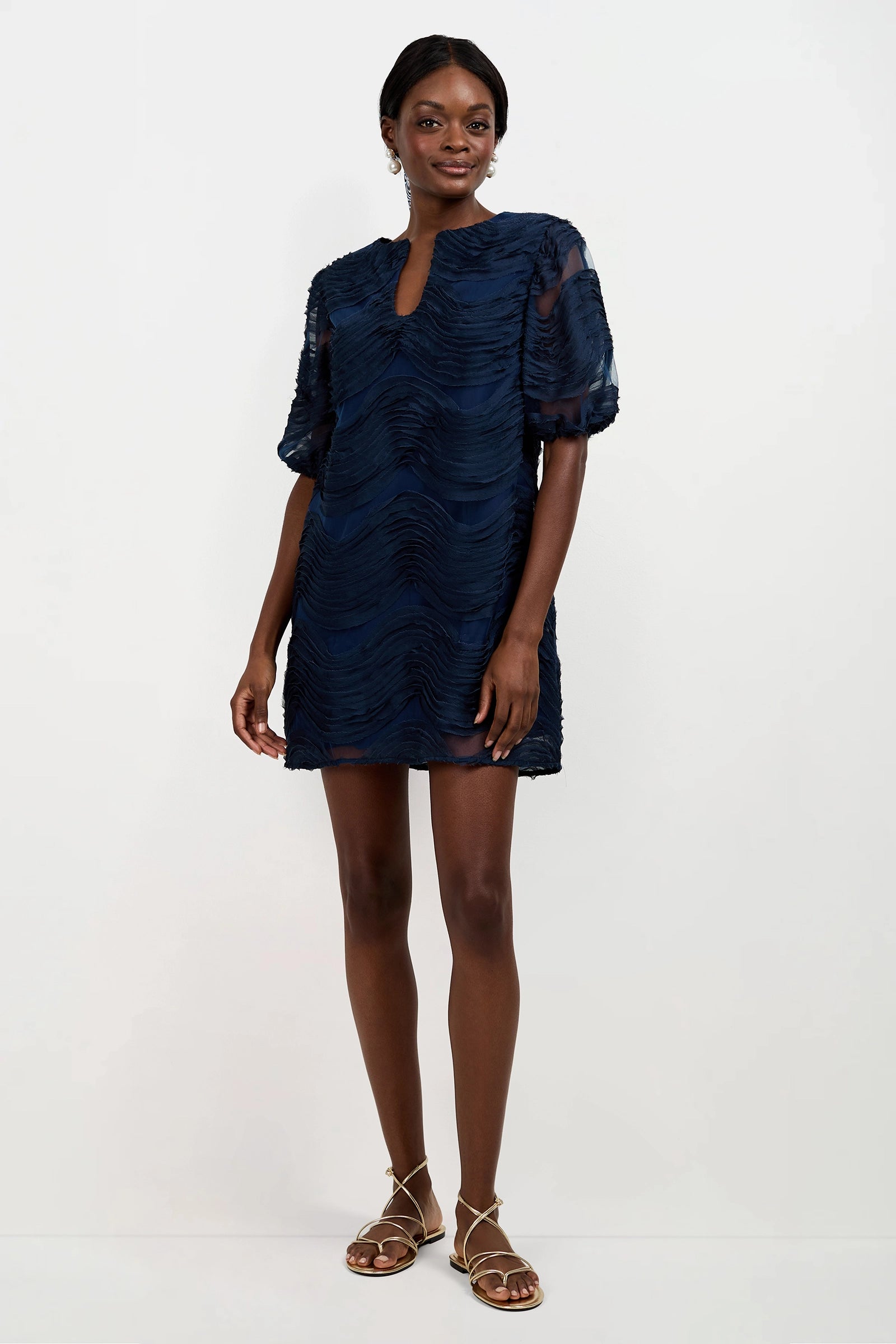 A woman poses against a white background wearing the Torchino Organza Mini Dress, a short textured navy blue piece with short sleeves and a keyhole neckline. She pairs it with sandals and smiles gently, one hand by her side, the other relaxed.
