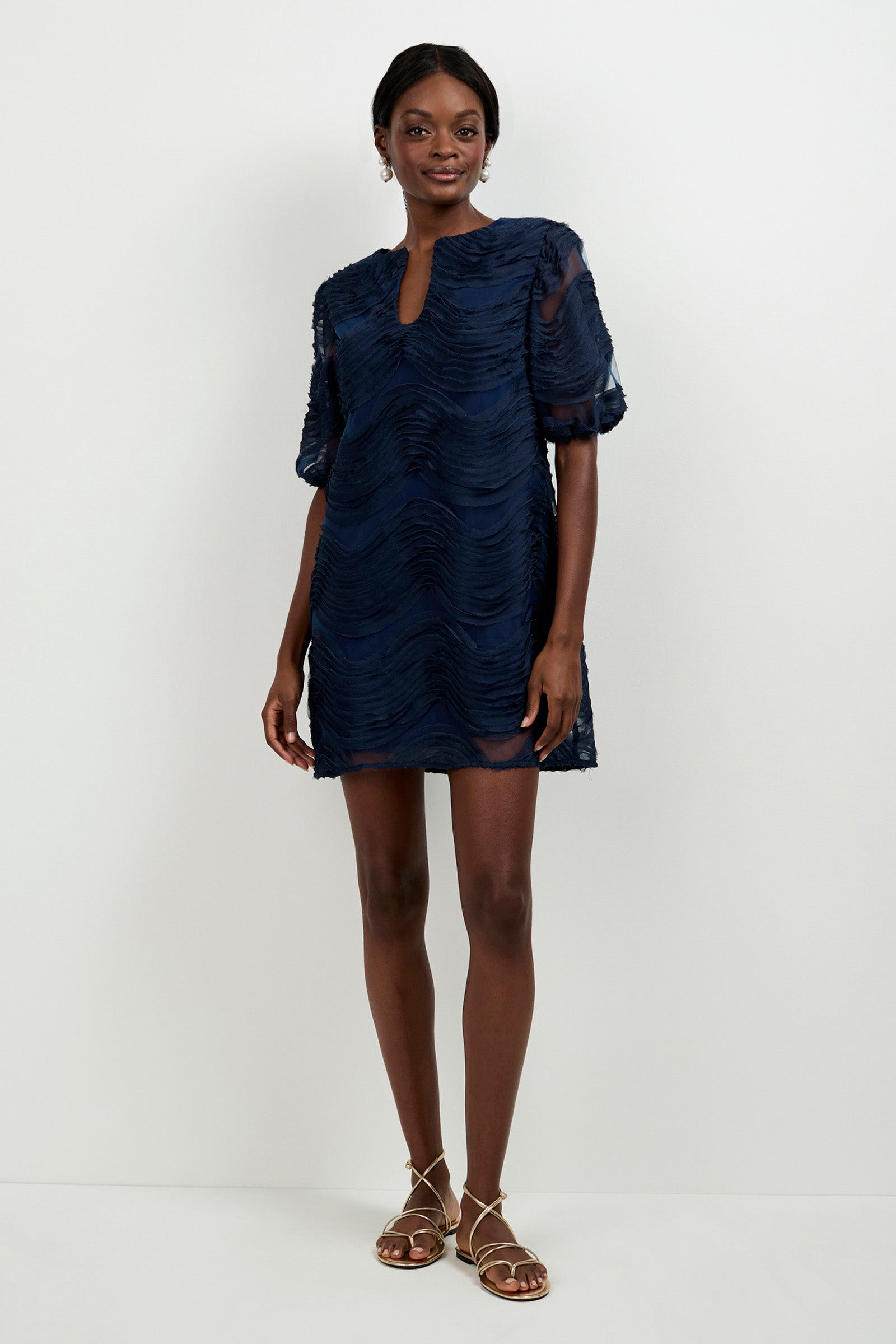 A woman stands against a white background wearing the Torchino Organza Mini Dress, which is navy blue, textured, short-sleeved with a keyhole neckline. She pairs it with gold strappy sandals and smiles slightly with her hands at her sides.