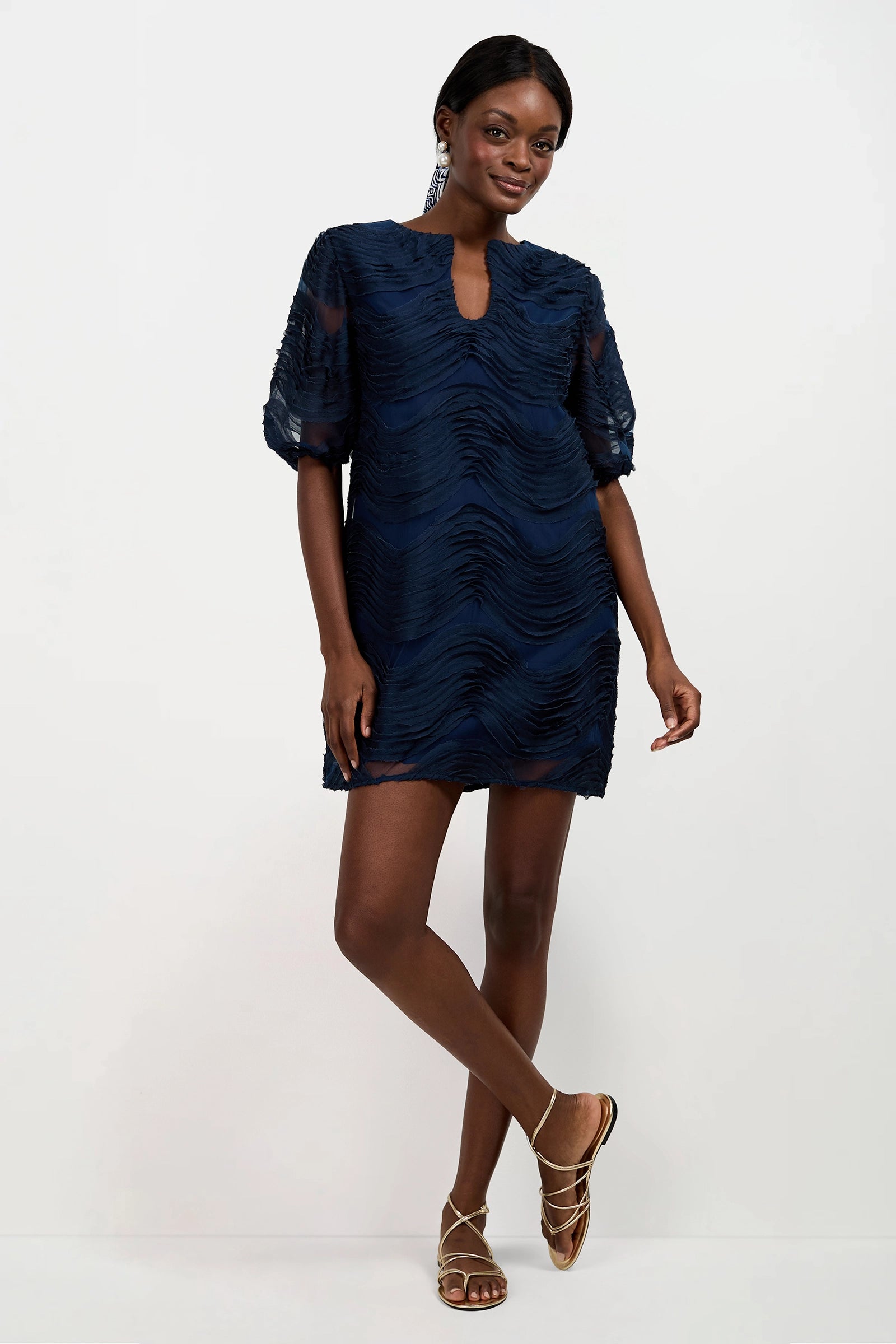 A woman wears the Torchino Organza Mini Dress—a short, textured navy blue dress with half sleeves and a notched neckline—paired with metallic strappy sandals and statement earrings, standing against a plain white background.