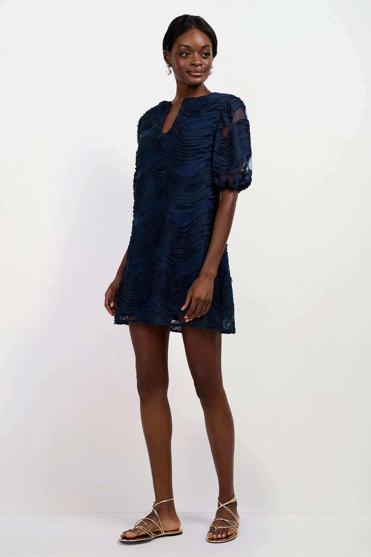 A woman stands against a plain white background wearing the Torchino Organza Mini Dress, a navy blue textured mini dress with short puff sleeves and a keyhole neckline, paired with gold strappy flat sandals.