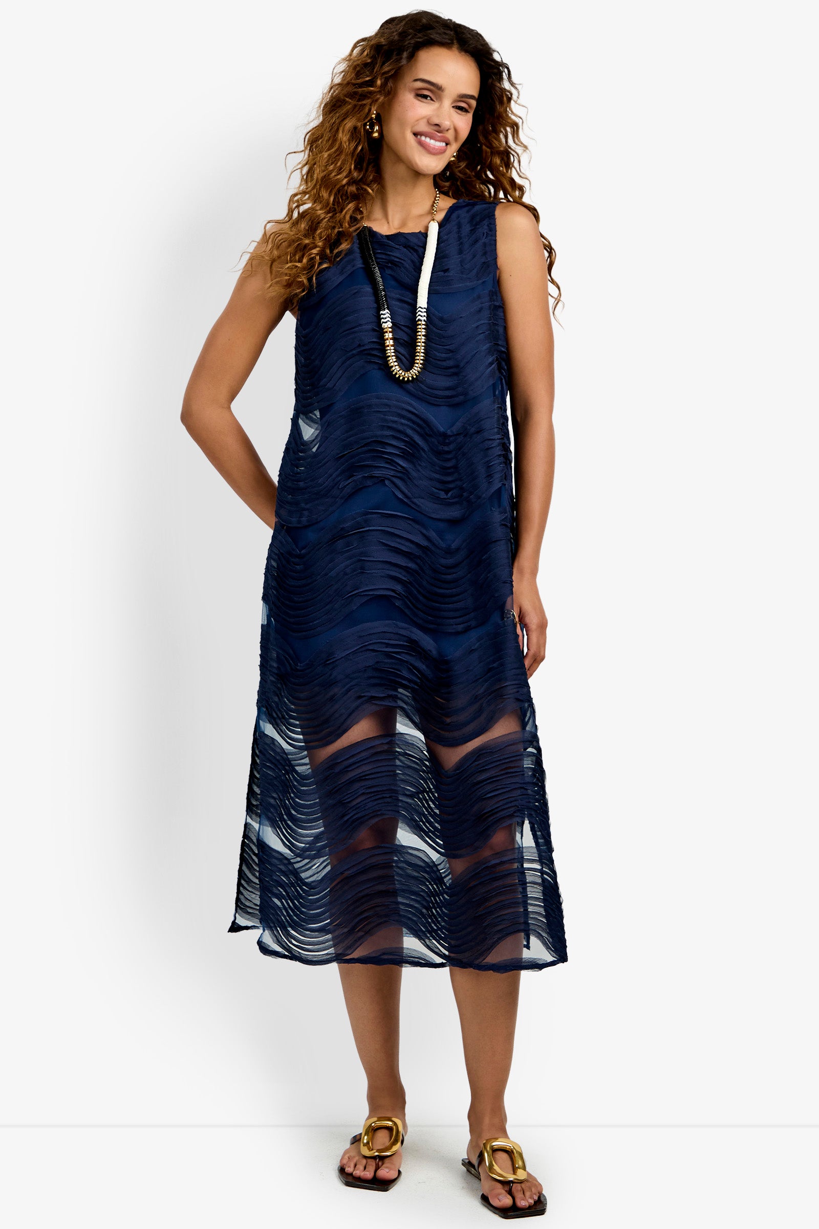 A woman stands and smiles against a white background, wearing the Belisama Organza Midi Dress—a sleeveless navy blue dress featuring textured wavy patterns and layered over a slip. She pairs the look with sandals, a long necklace, and has curly hair.