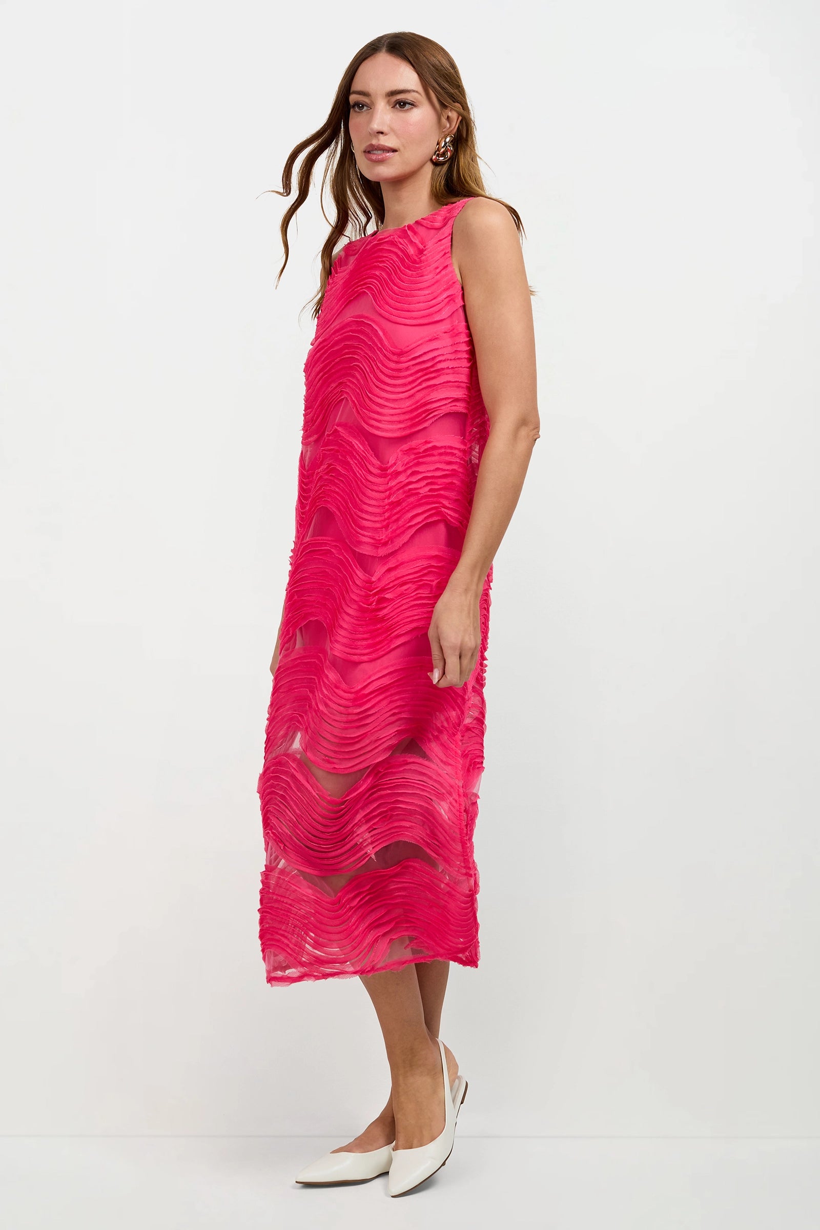 A woman stands against a white background wearing the Belisama Organza Midi Dress, a sleeveless, bright pink, mid-calf dress with textured wavy patterns, paired with white pointed-toe heels.