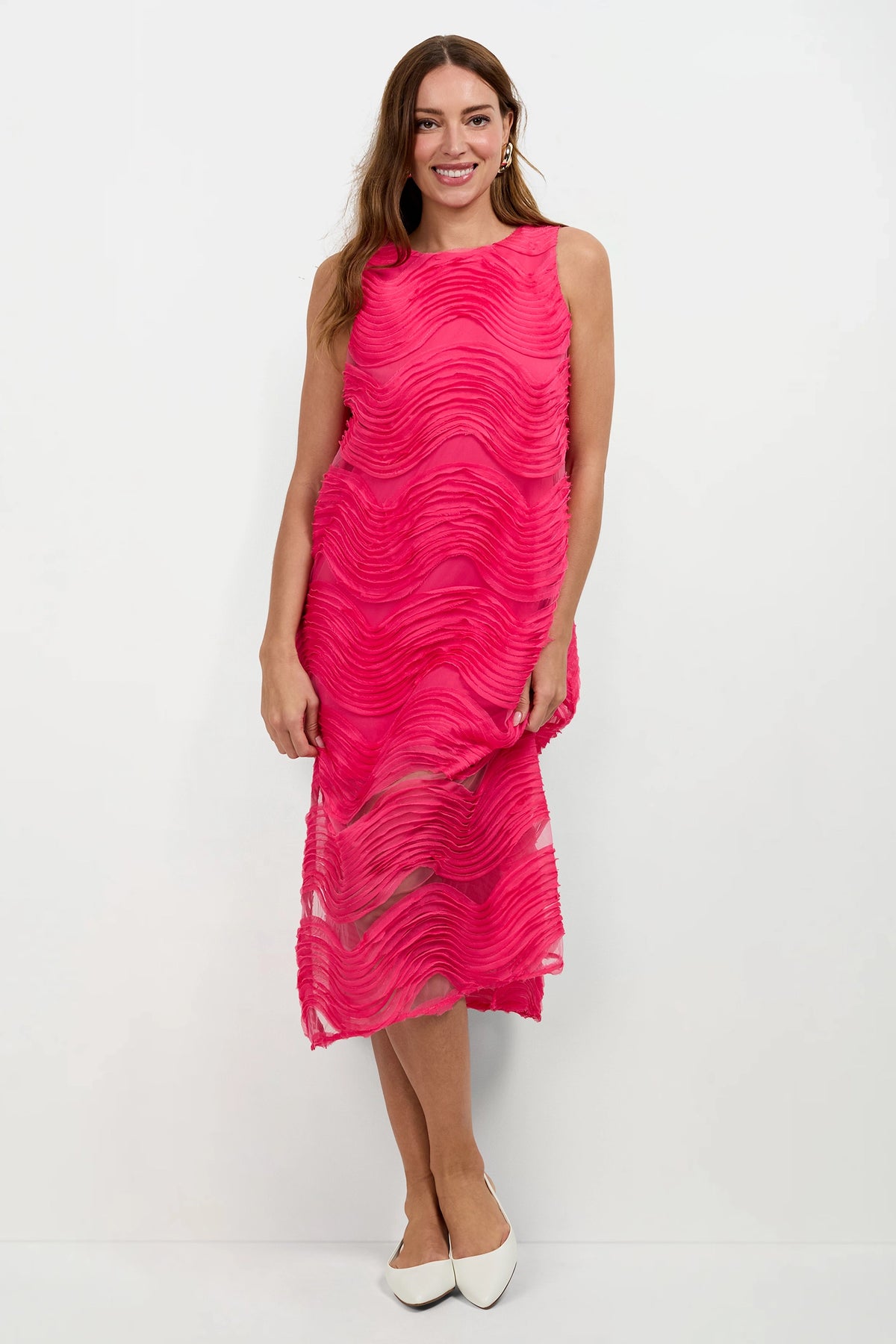 A woman with long brown hair smiles while wearing the Belisama Organza Midi Dress—a sleeveless, bright pink textured midi dress with wavy patterns—paired with white pointed-toe flats against a plain white background.