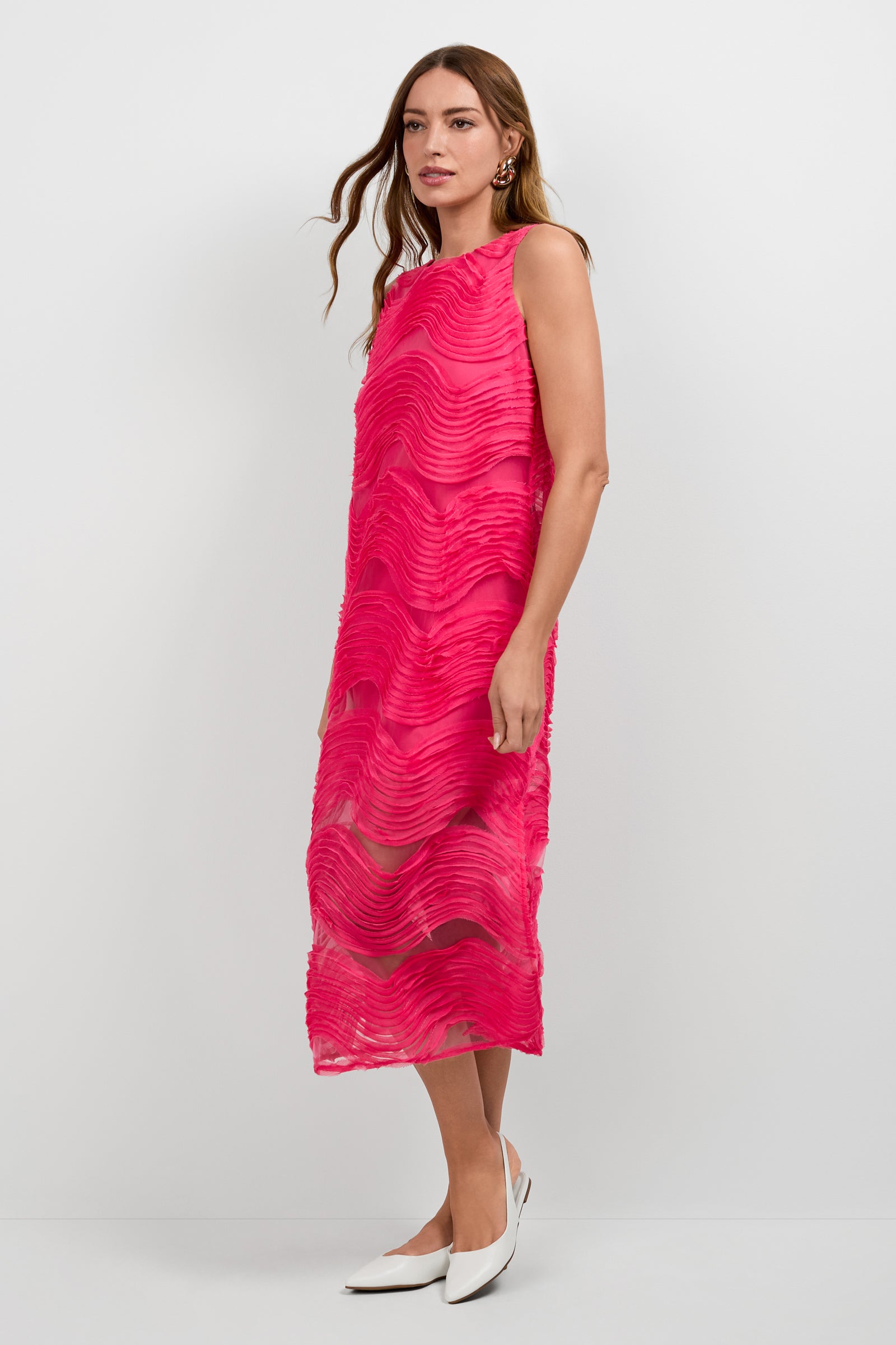 A woman stands against a white background wearing the sleeveless, bright pink Belisama Organza Midi Dress with textured wave patterns and white pointed-toe shoes.