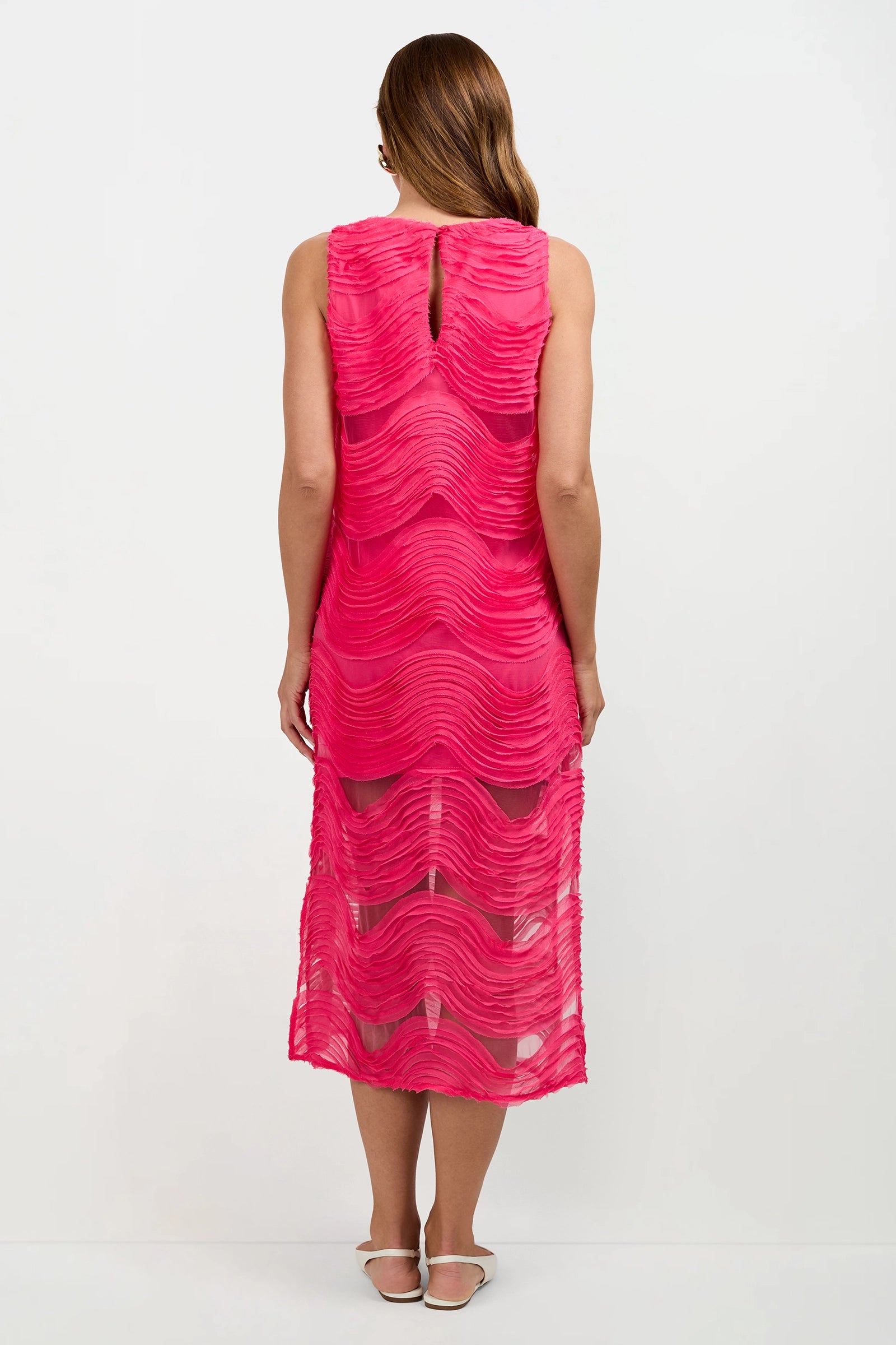A person with long brown hair, seen from the back, wears the Belisama Organza Midi Dress—a sleeveless, knee-length bright pink dress with wavy textured layers and a sheer lower skirt—paired with white flat shoes.