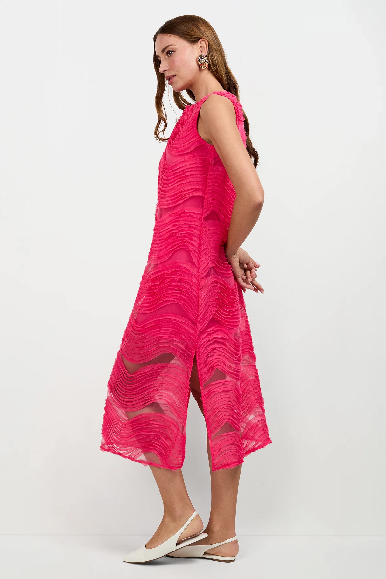 A woman with long brown hair stands sideways against a plain background, wearing the sleeveless Belisama Organza Midi Dress in bright pink with side slits, styled with white pointed-toe slingback shoes and large earrings.