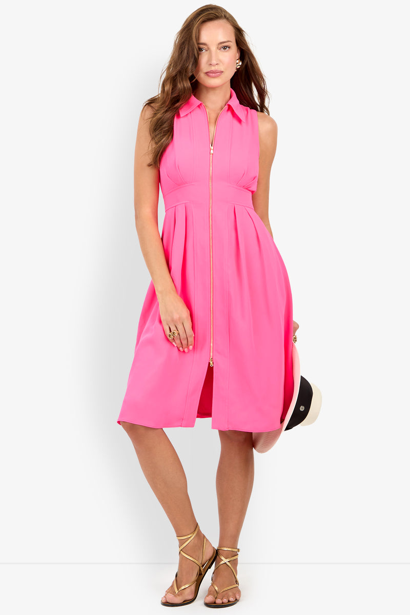 Woman in the Farfalla Sleeveless Midi Dress, a bright pink, knee-length dress with a front zipper, holding a hat and wearing gold sandals against a plain white background.
