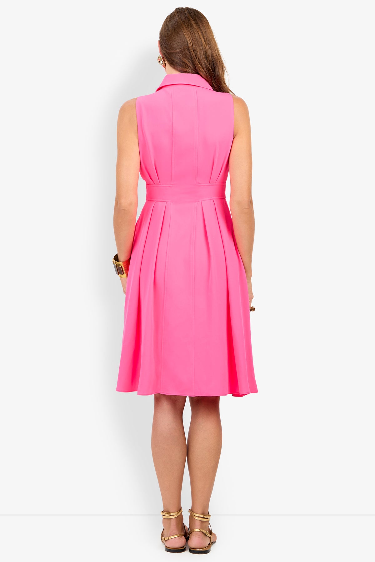 A woman with long brown hair and gold sandals wears the Farfalla Sleeveless Midi Dress, a bright pink, knee-length style featuring a fitted waist and pleated skirt.