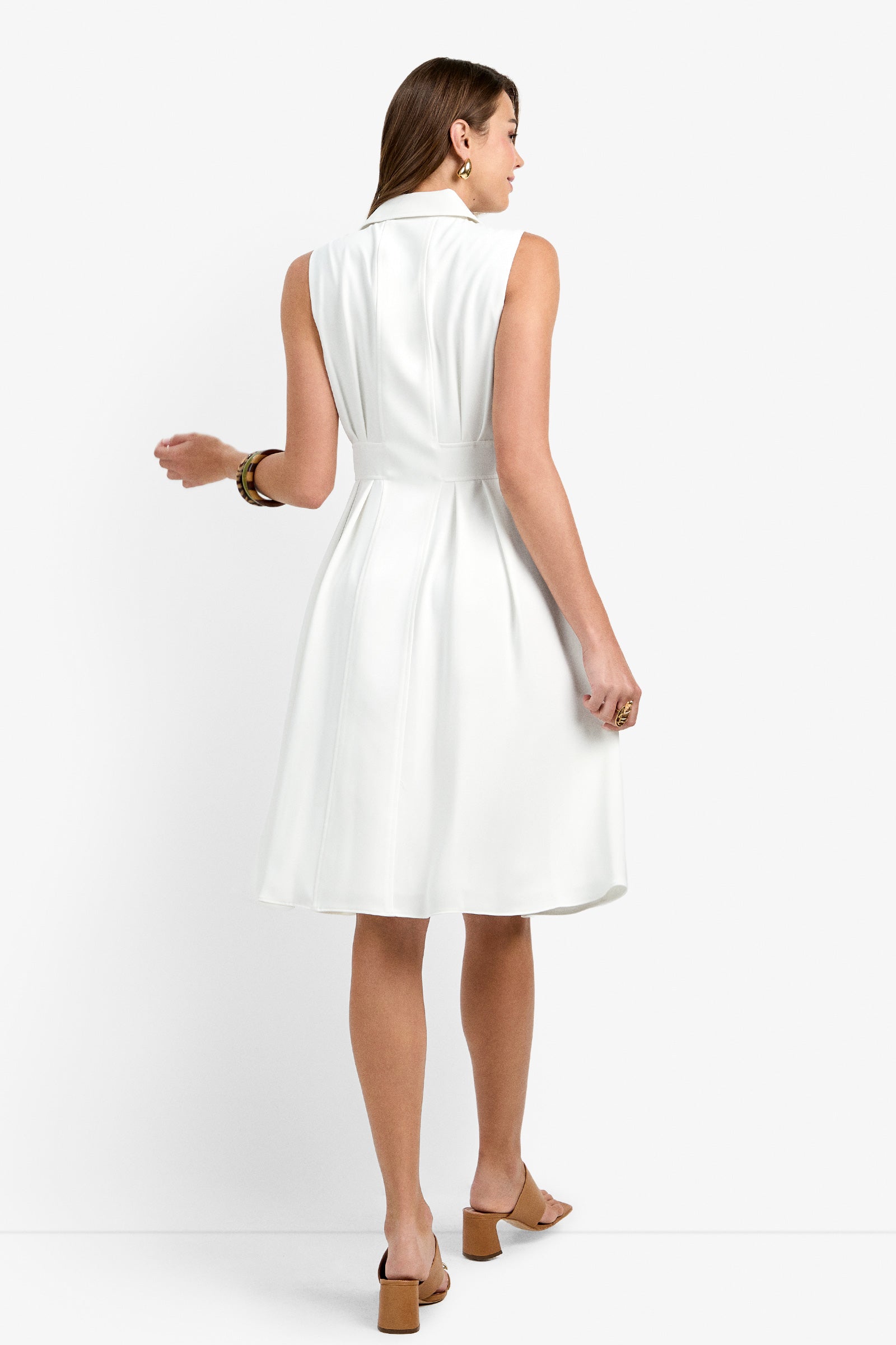 Woman with brown hair stands with her back to the camera, wearing the Farfalla Sleeveless Midi Dress—white, fitted at the waist with a flared knee-length skirt—paired with tan heeled sandals, gold earrings, and a bracelet.