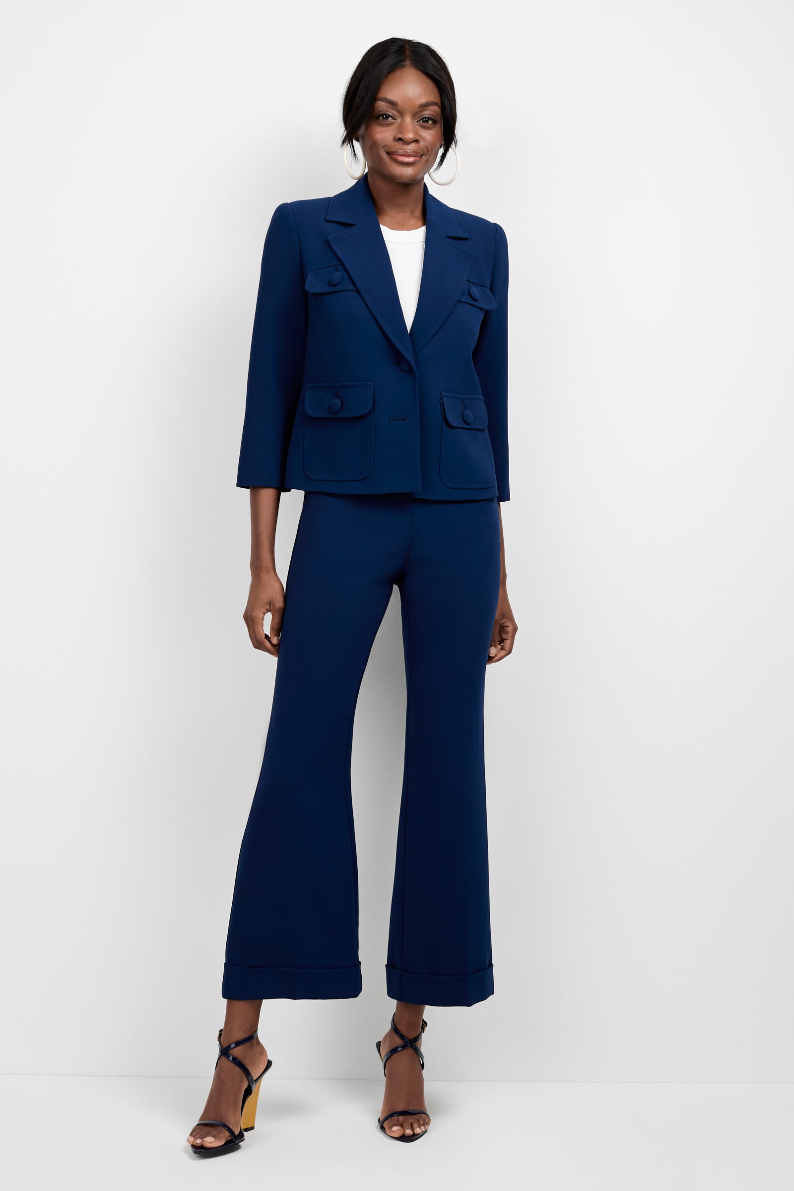 A woman smiles, standing against a white background wearing the Bicocca Patch Pocket Jacket in navy blue with matching wide-leg pants, a white top, and black high-heeled sandals; one hand rests by her side.