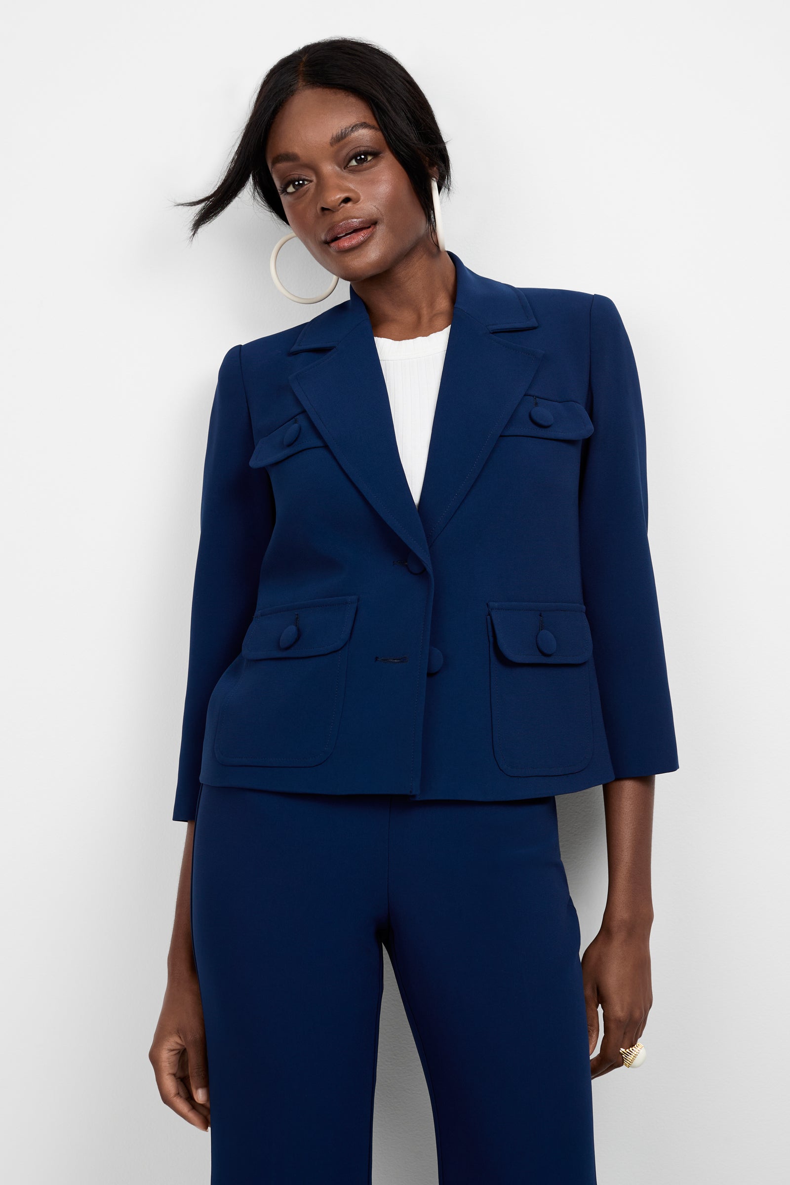 A woman wears the Bicocca Patch Pocket Jacket—a tailored navy blue suit jacket with front flap pockets and three-quarter sleeves—over a white top, paired with short styled hair and large hoop earrings, against a plain white background.