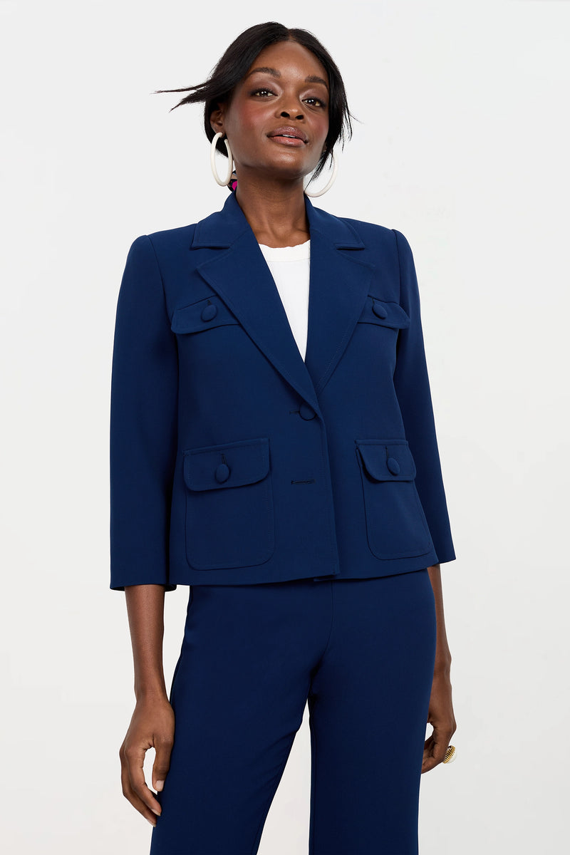A woman stands confidently in the Bicocca Patch Pocket Jacket, a dark blue tailored blazer with front pockets, paired with matching pants, a white top, and large hoop earrings against a plain light background.