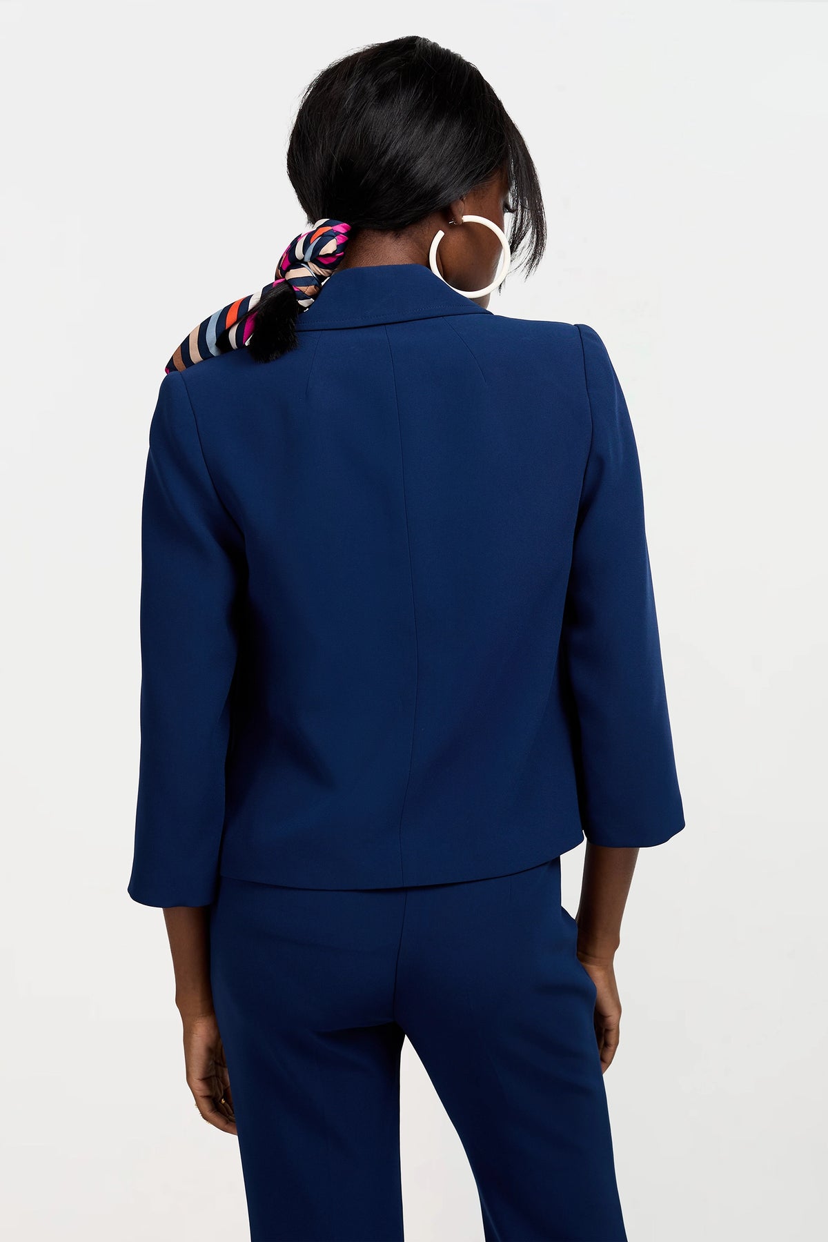 A woman is seen from behind wearing the Bicocca Patch Pocket Jacket in dark blue with three-quarter sleeves, a colorful striped scarf tied around her hair, and large hoop earrings, set against a plain white background.