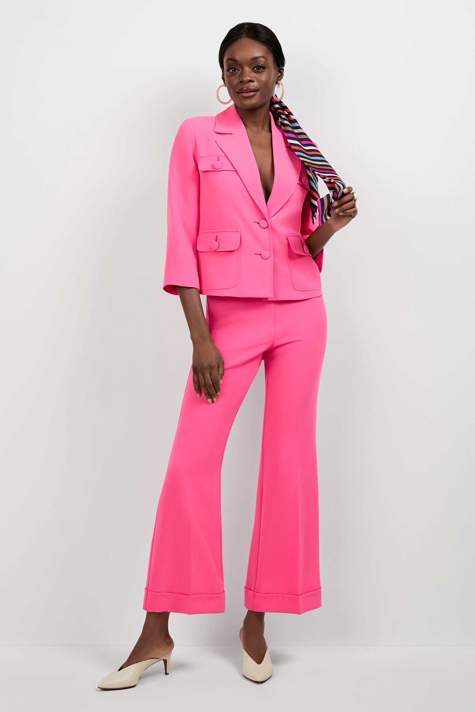 A woman stands against a white background wearing the Bicocca Patch Pocket Jacket in bright pink with wide-legged pants. She accessorizes with a colorful striped hair scarf and white pointed-toe heels.