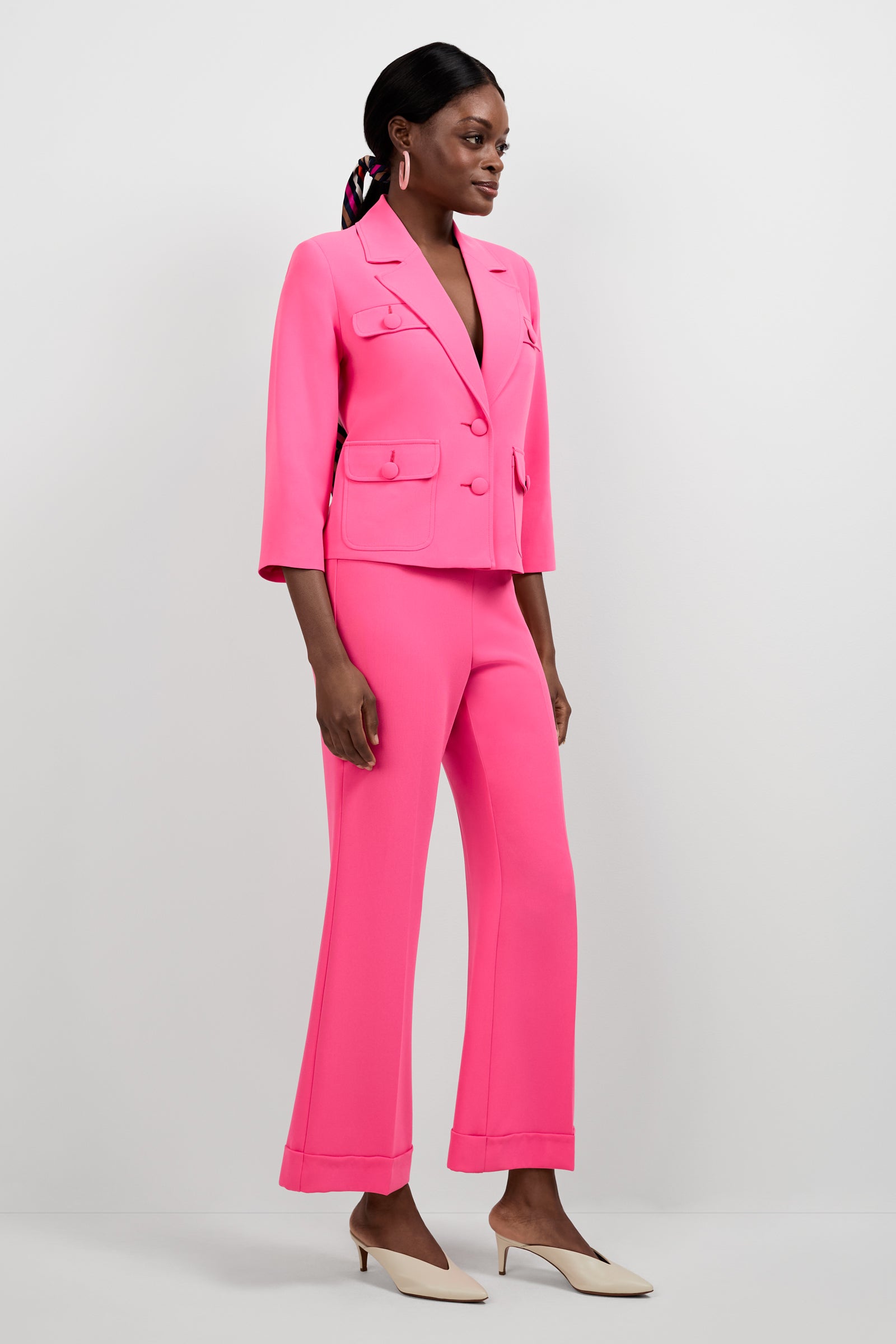 A woman stands against a plain background wearing the Bicocca Patch Pocket Jacket in bright pink with matching flared pants and white low-heeled shoes. Her hair is tied back with a scarf, and she looks slightly to the side.