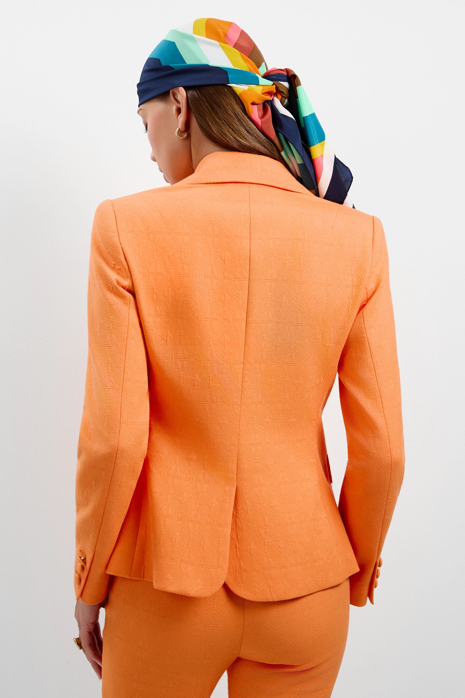 A woman faces away in the Incanta Fitted Blazer, styled with a colorful striped headscarf against a plain white background.